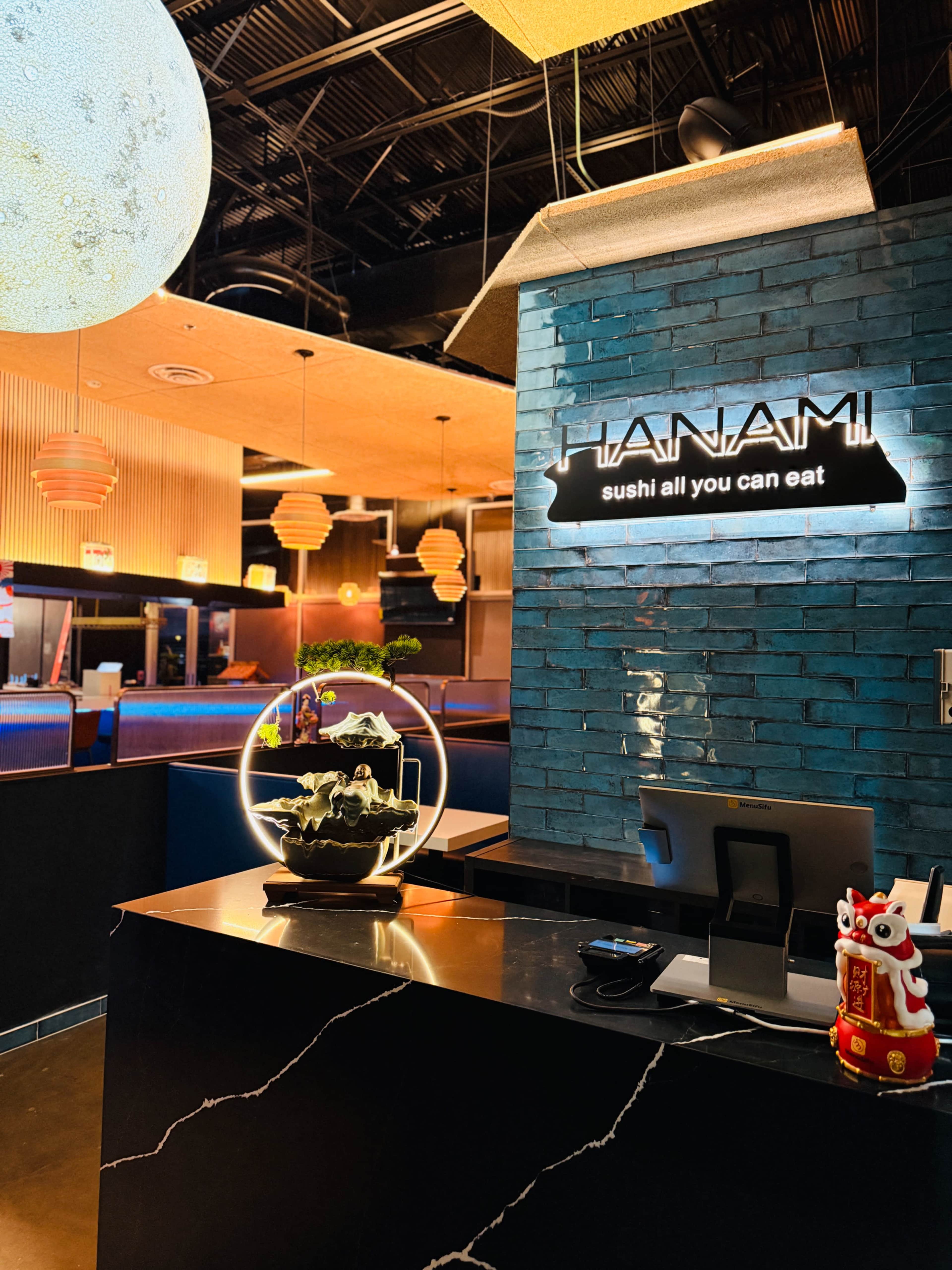 The interior of a sushi restaurant features a modern design with decorative lighting, a reception area, and a unique display with a miniature landscape.