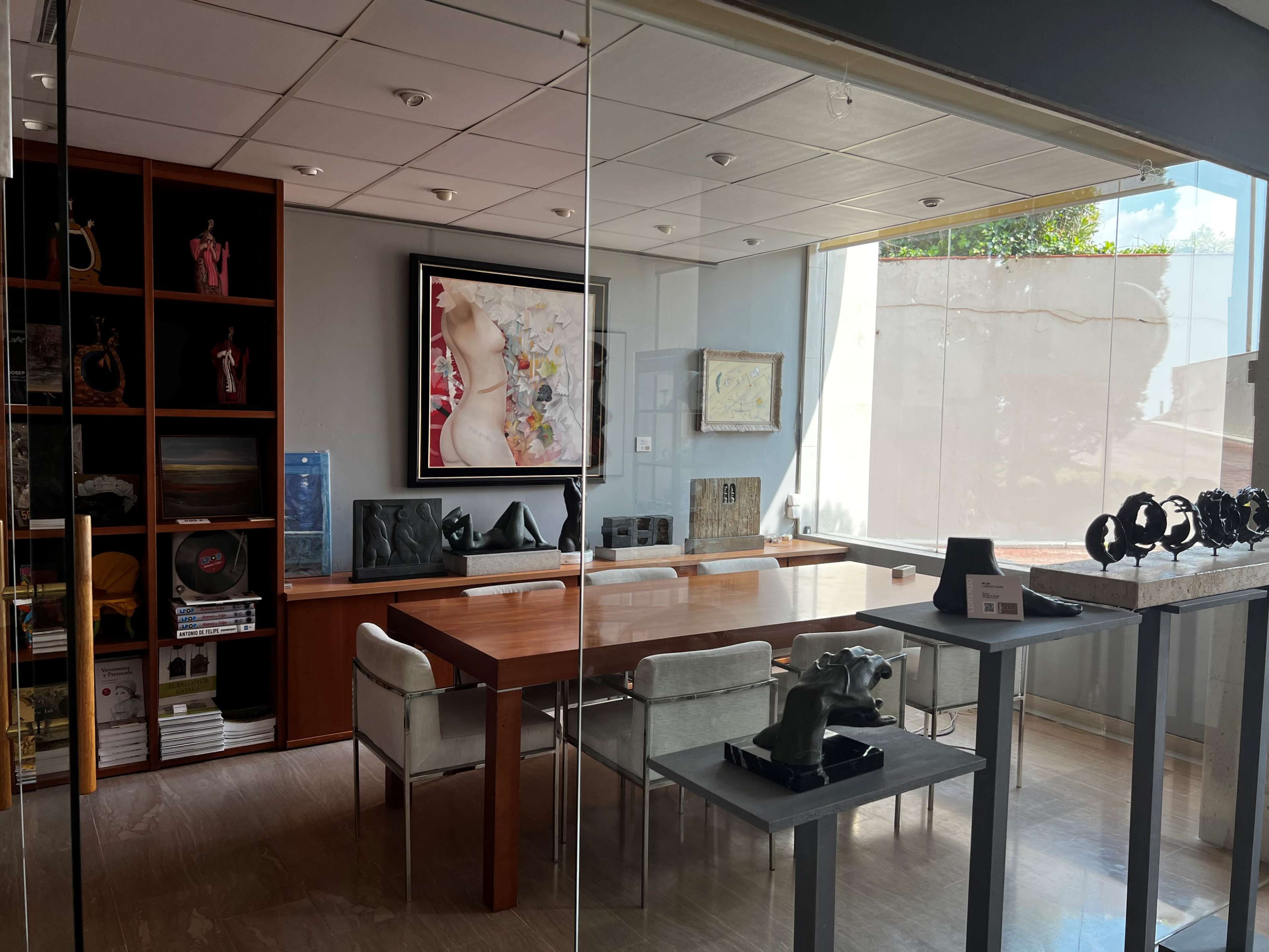 The image shows a modern office space featuring a wooden table surrounded by chairs, a wall display of art and sculptures, and large windows allowing natural light to enter.