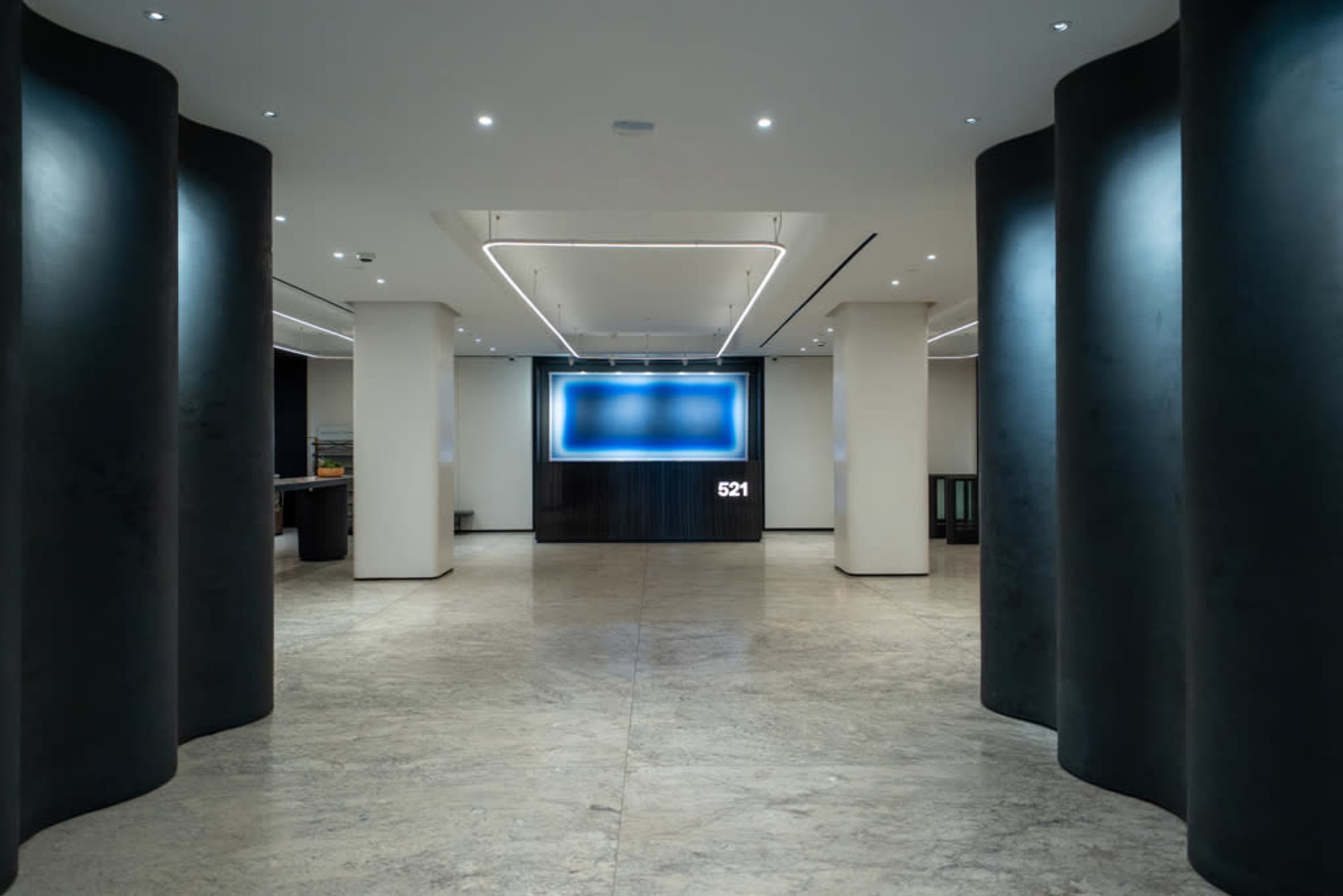 The image shows a spacious lobby area with smooth stone flooring, illuminated by recessed lighting, flanked by large, dark columns, and featuring a backlit wall display.