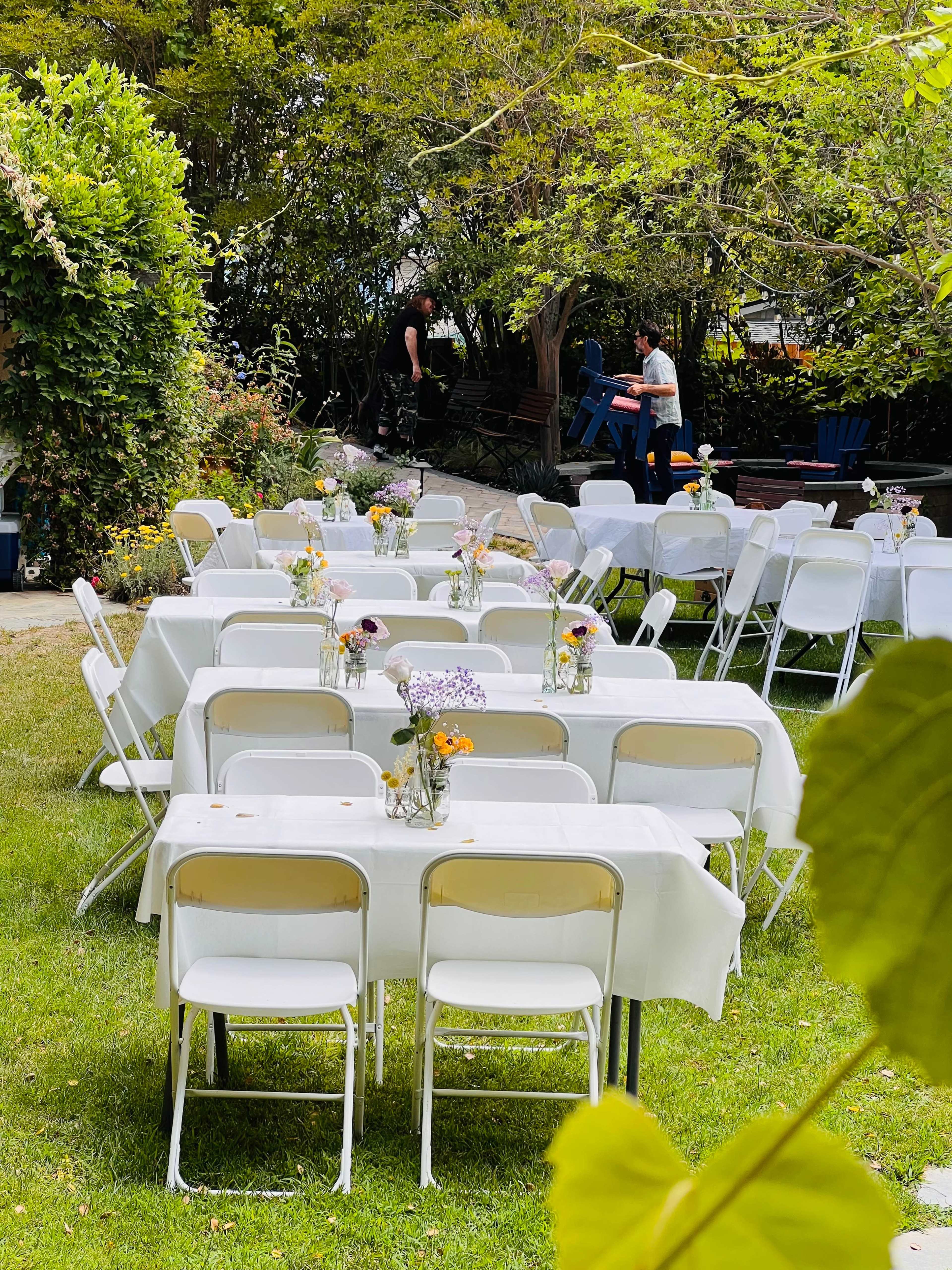 A row of neatly arranged white tables and chairs is set up in a garden, with small vases of flowers on each table.