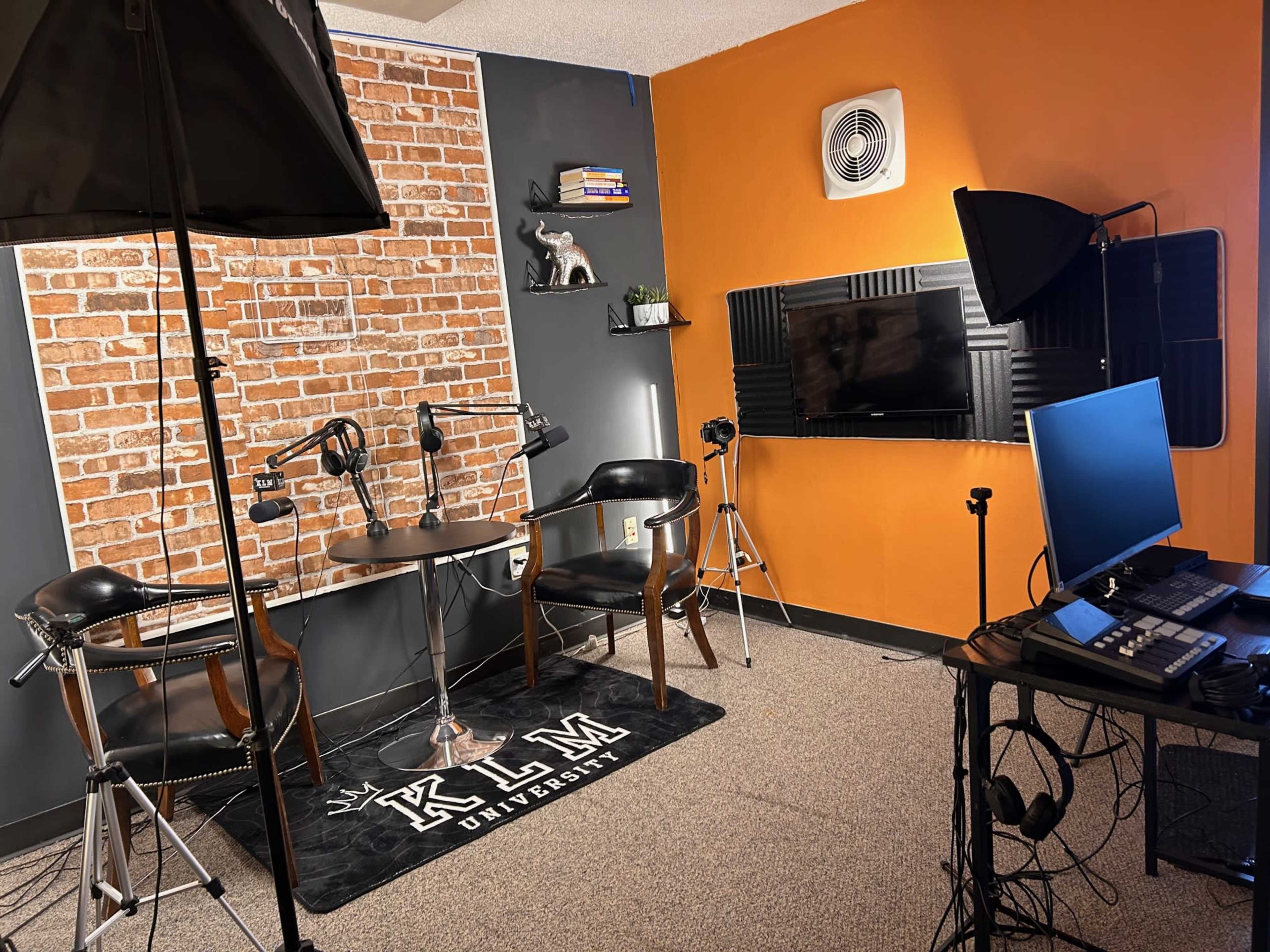 A small studio setup with two chairs and a table against a brick wall, a television mounted on a black foam panel, and lighting equipment nearby.
