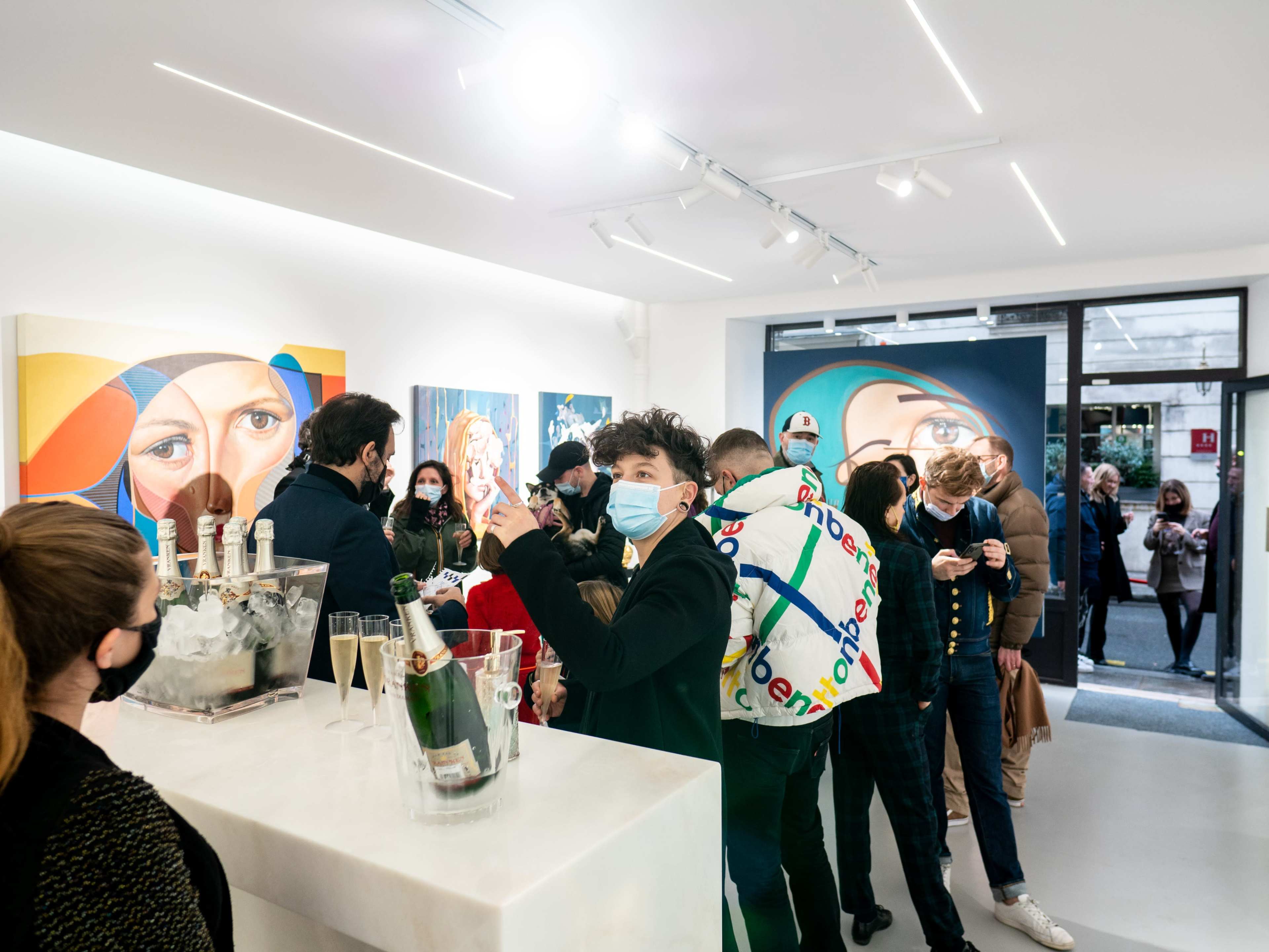A crowd gathers in a modern art gallery, enjoying drinks while admiring colorful portraits on the walls.