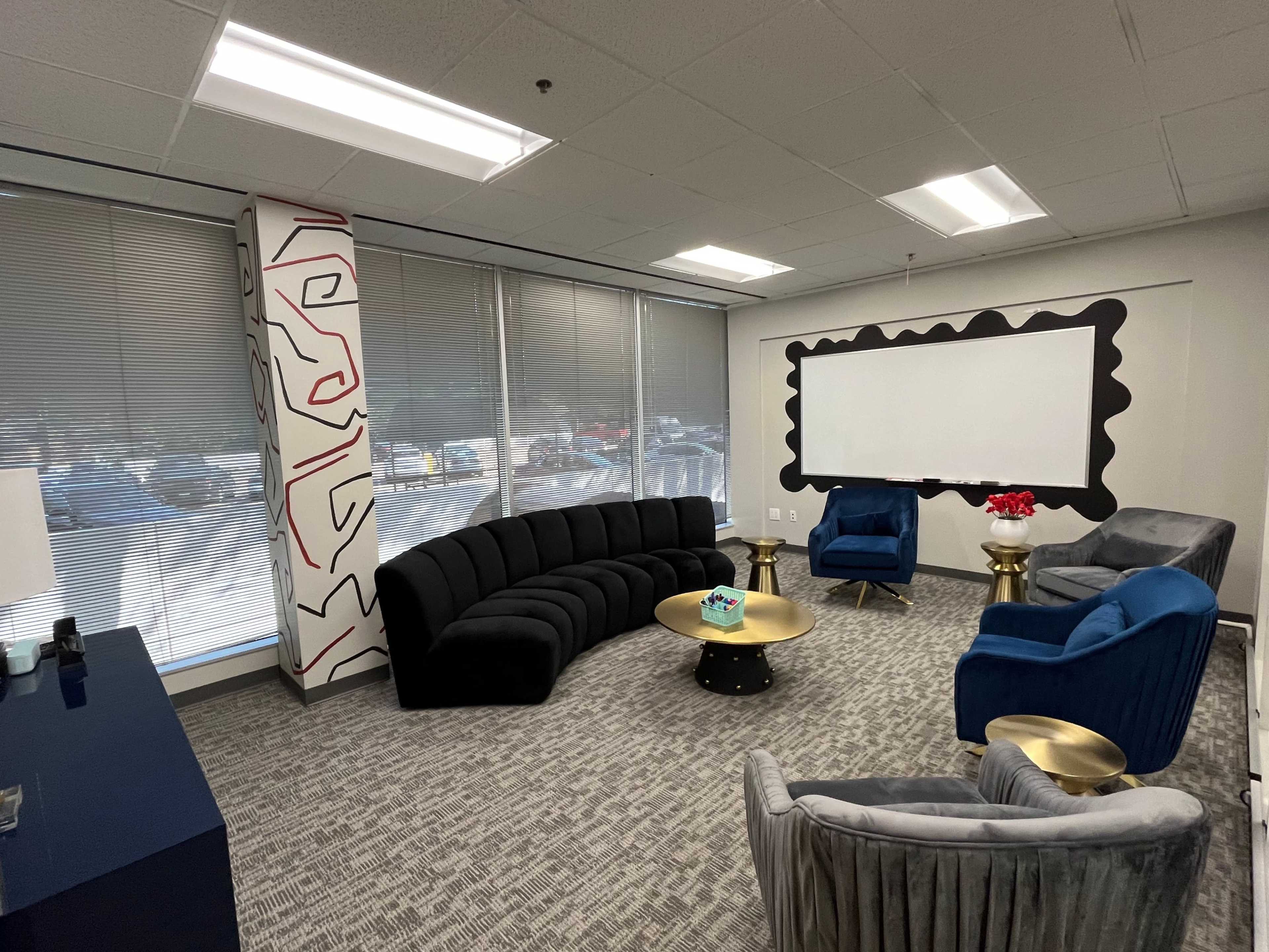 A modern office lounge features a curved black sofa, blue chairs, a round gold coffee table, and a large whiteboard against a wall.