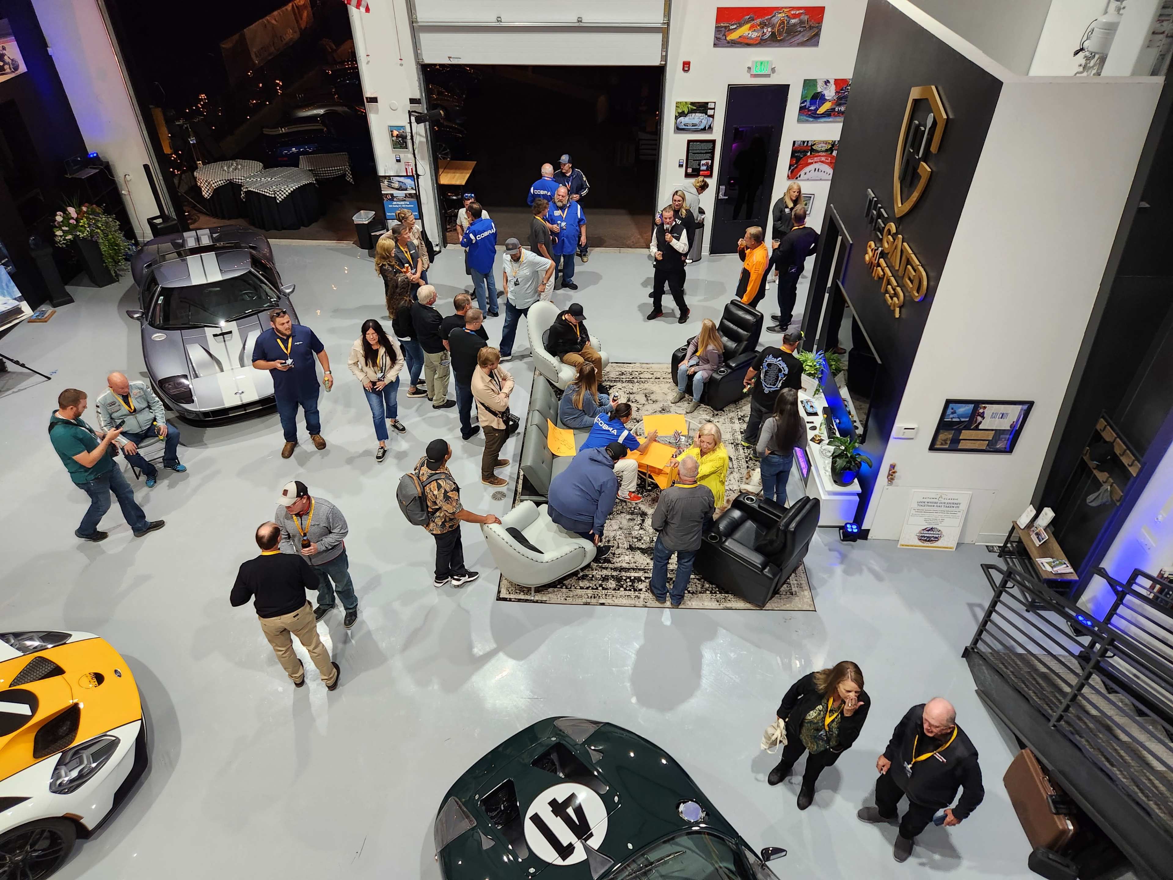 A group of people is gathered in a spacious showroom featuring luxury cars, with some engaging in conversation while others network and socialize.