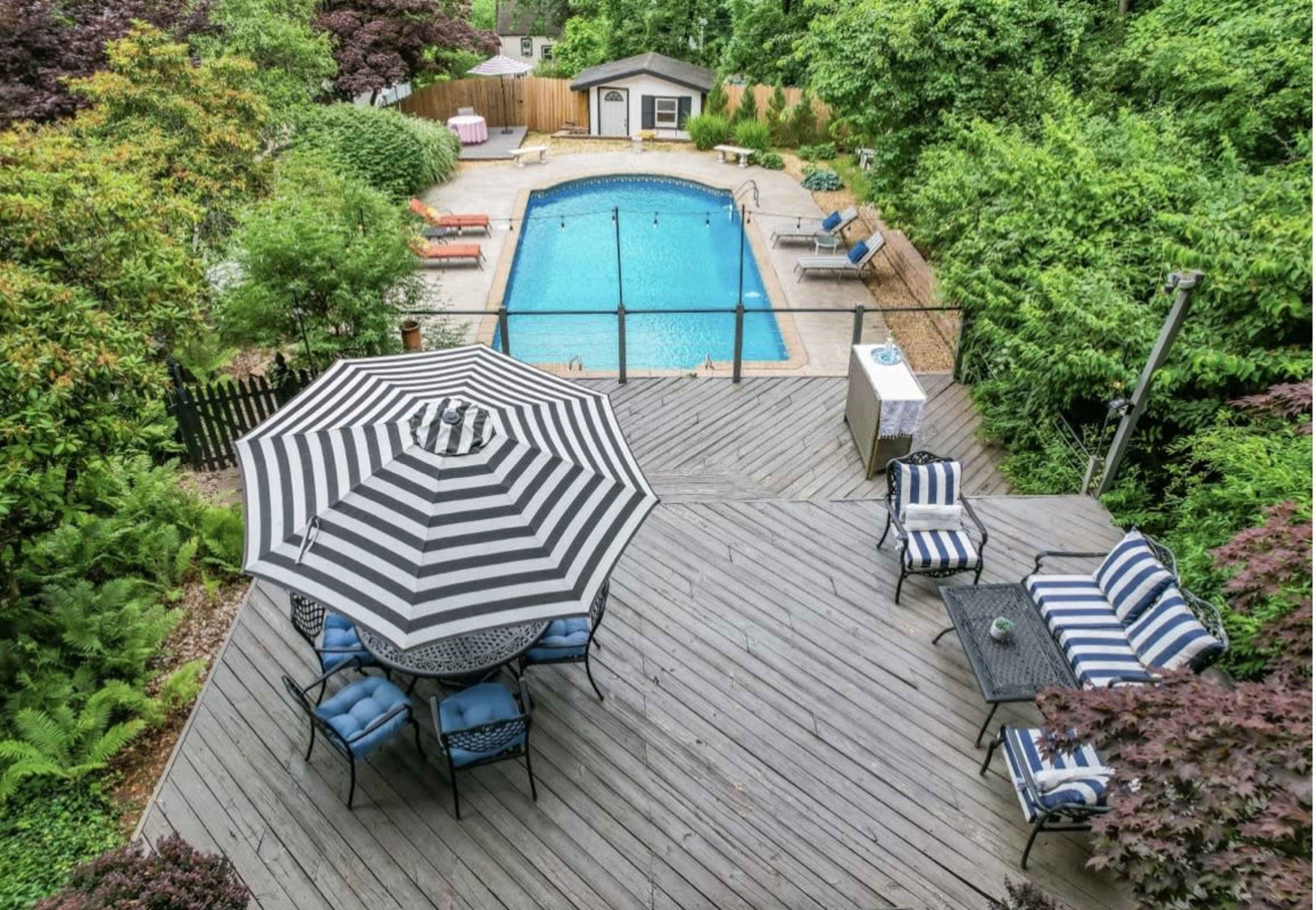 The image shows a backyard with a swimming pool surrounded by a wooden deck, patio furniture, and lush green trees.