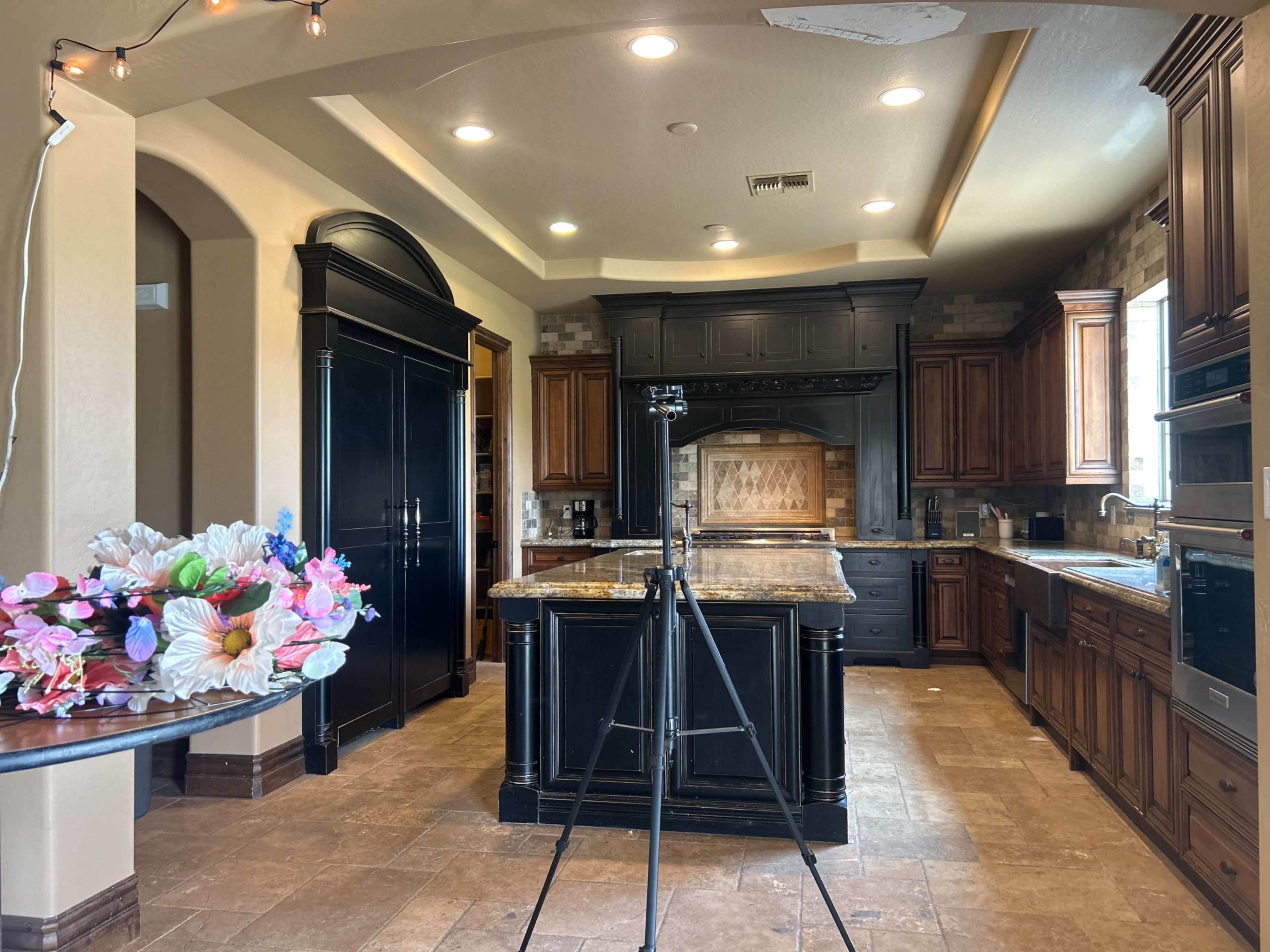 A modern kitchen features dark wood cabinetry, a large central island, and various appliances arranged around the space, with a camera tripod set up on one side and a floral arrangement on a table.