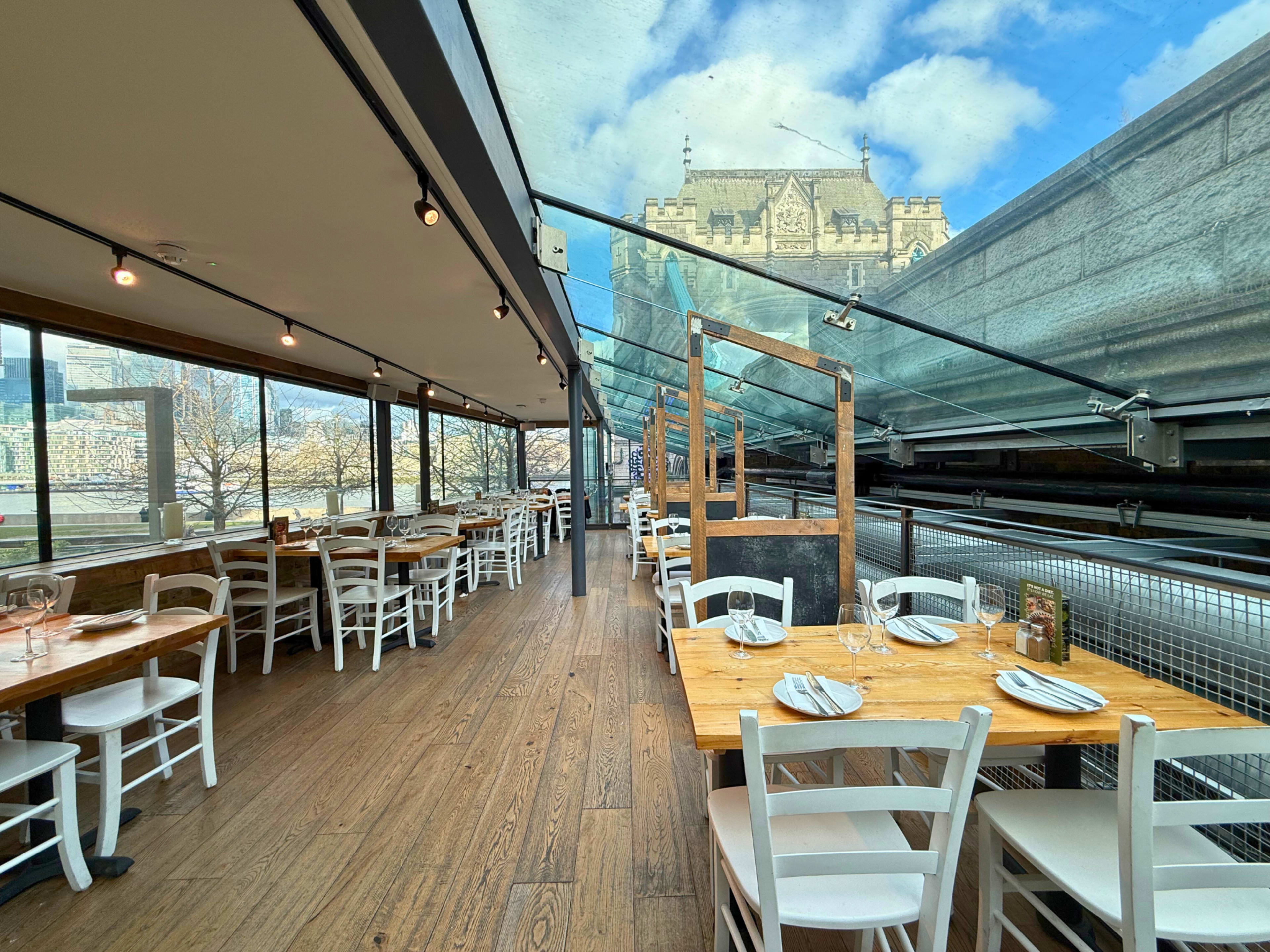 The image shows an indoor dining area with wooden tables and white chairs, featuring large windows that offer a view of a building outside.