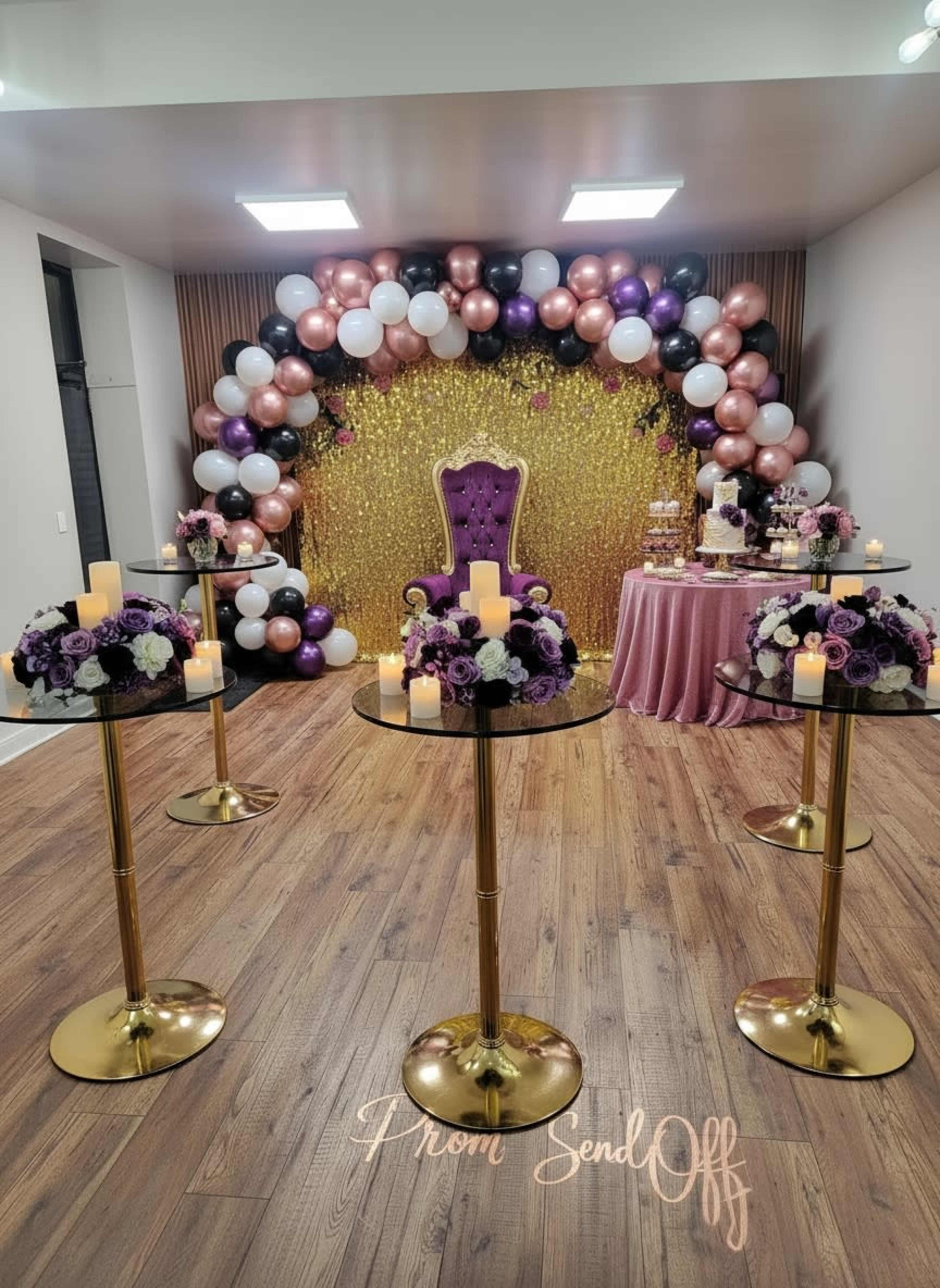 The image shows a decorative event space with a gold backdrop, a purple and gold throne chair, tables adorned with floral arrangements, and a balloon arch in black, white, and pink colors.