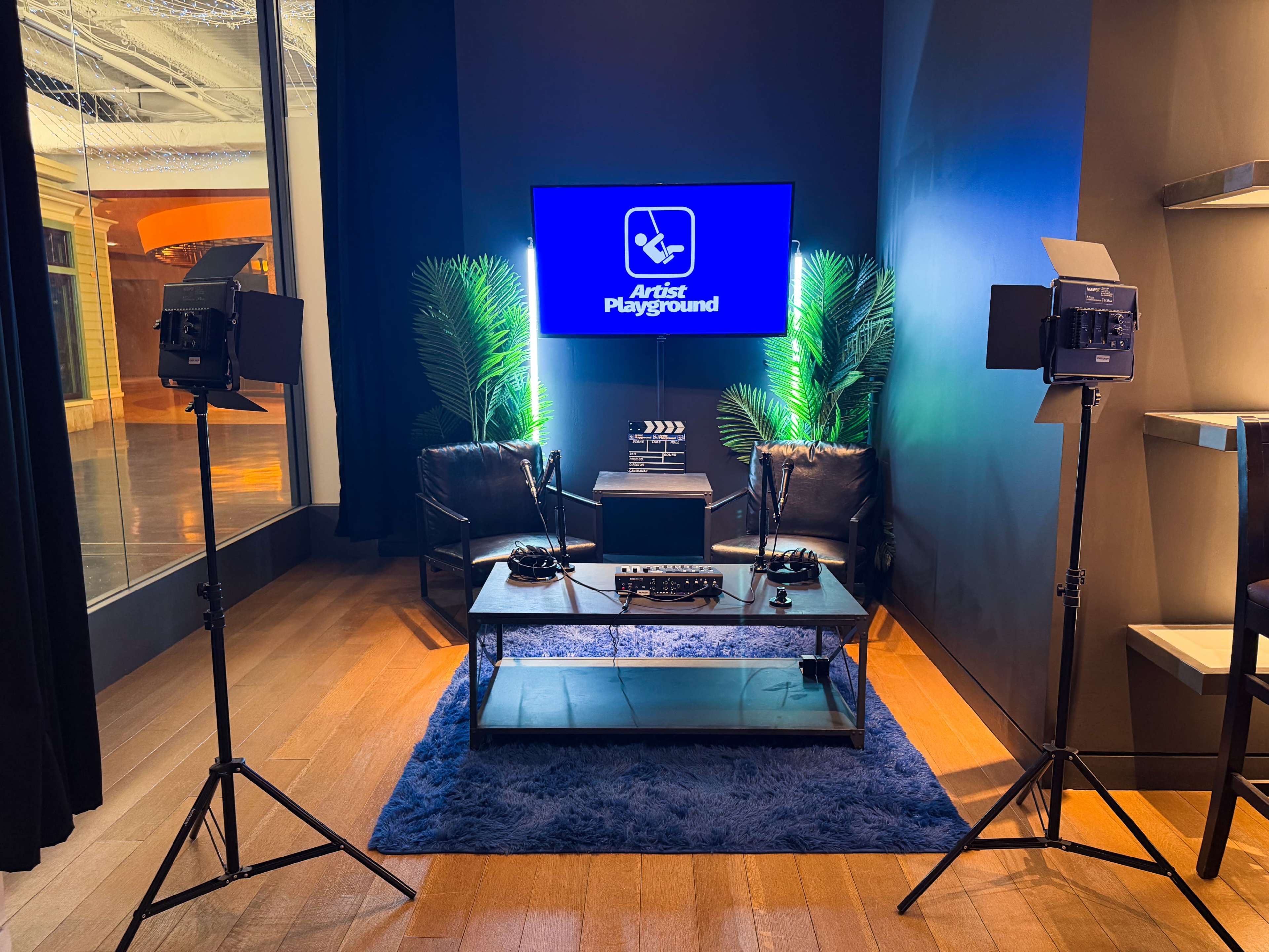 The image shows a small studio setup with two video lights, a coffee table, and two chairs, all featuring a large screen displaying the logo for "Artist Playground" in a modern space.