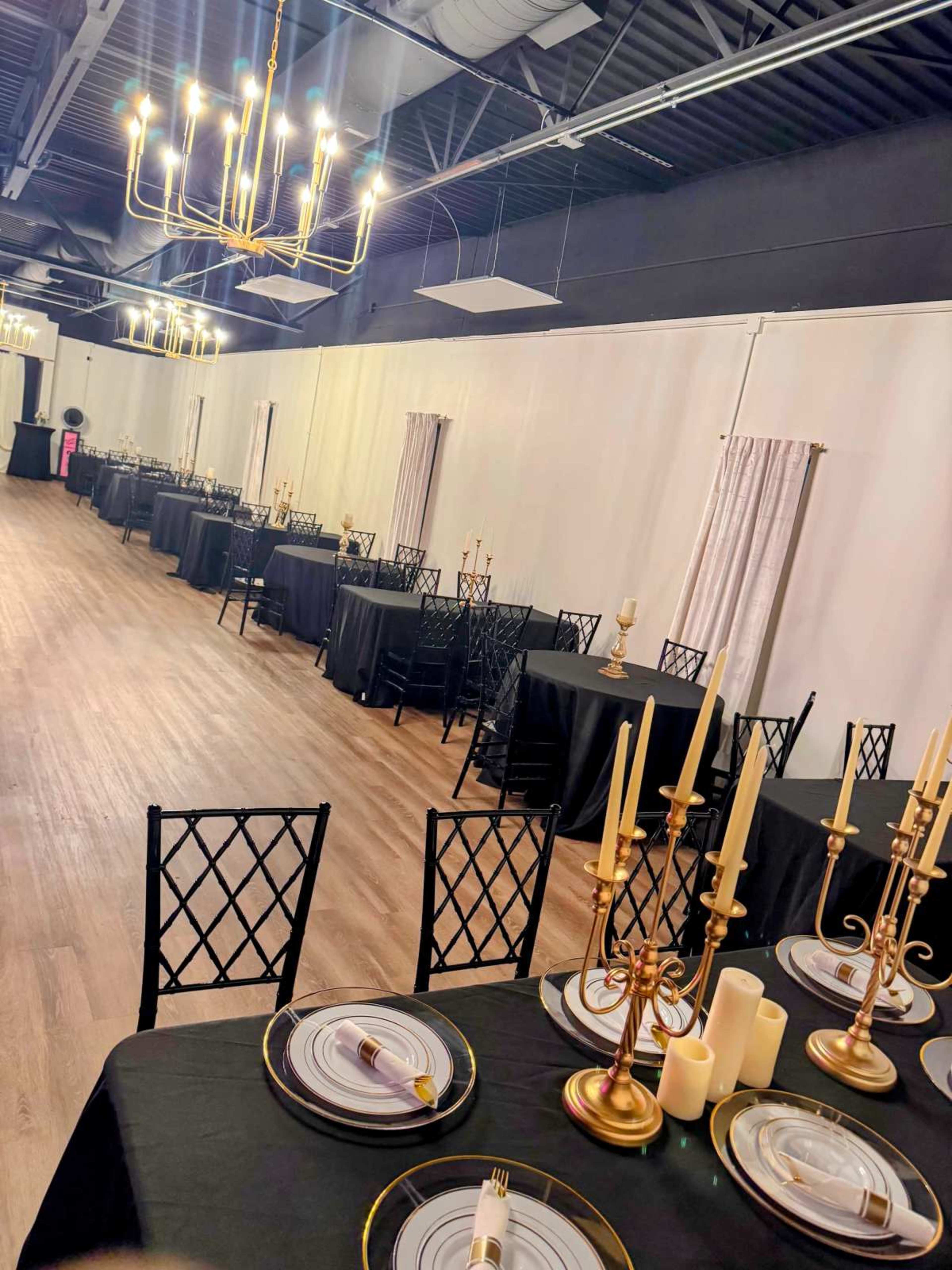 A long banquet hall is set with black tables, gold candlesticks, and white candles, illuminated by a chandelier overhead.