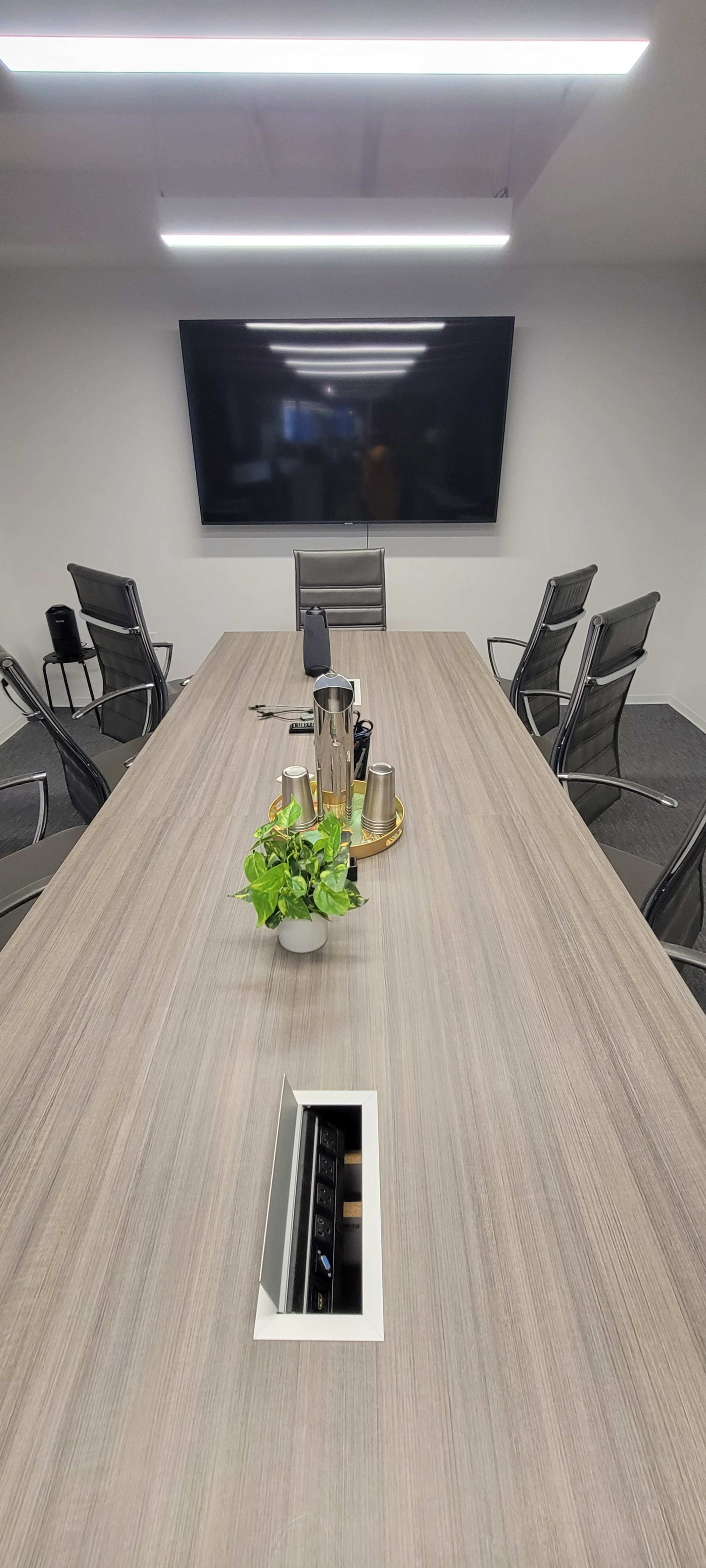 A modern conference room features a long wooden table with a plant centerpiece, surrounded by several black chairs, and a large screen on the wall.