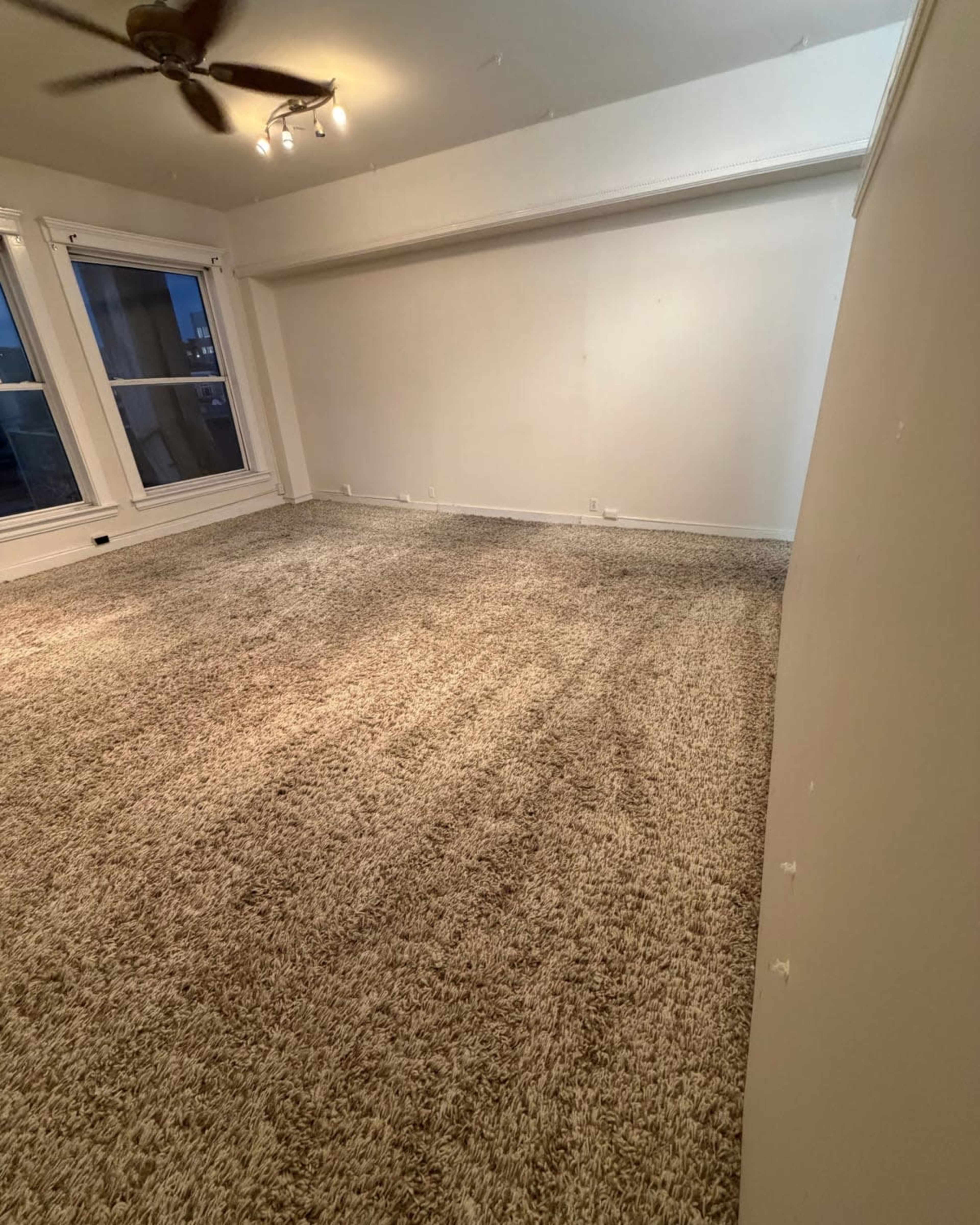 The image shows an empty room with beige carpet, large windows, and a ceiling fan.