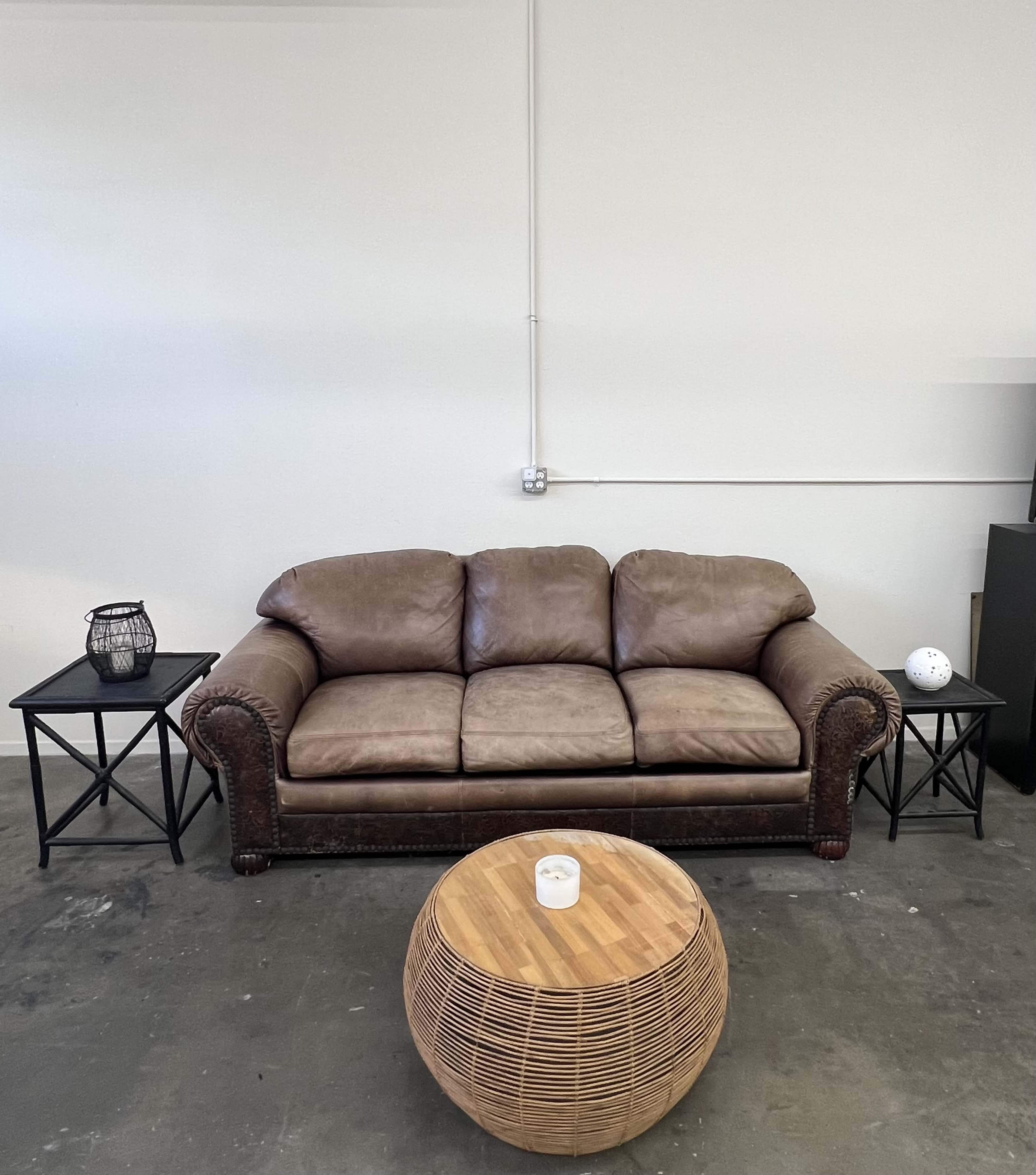 The image shows a brown leather couch flanked by two black side tables, with a round woven coffee table in the center.