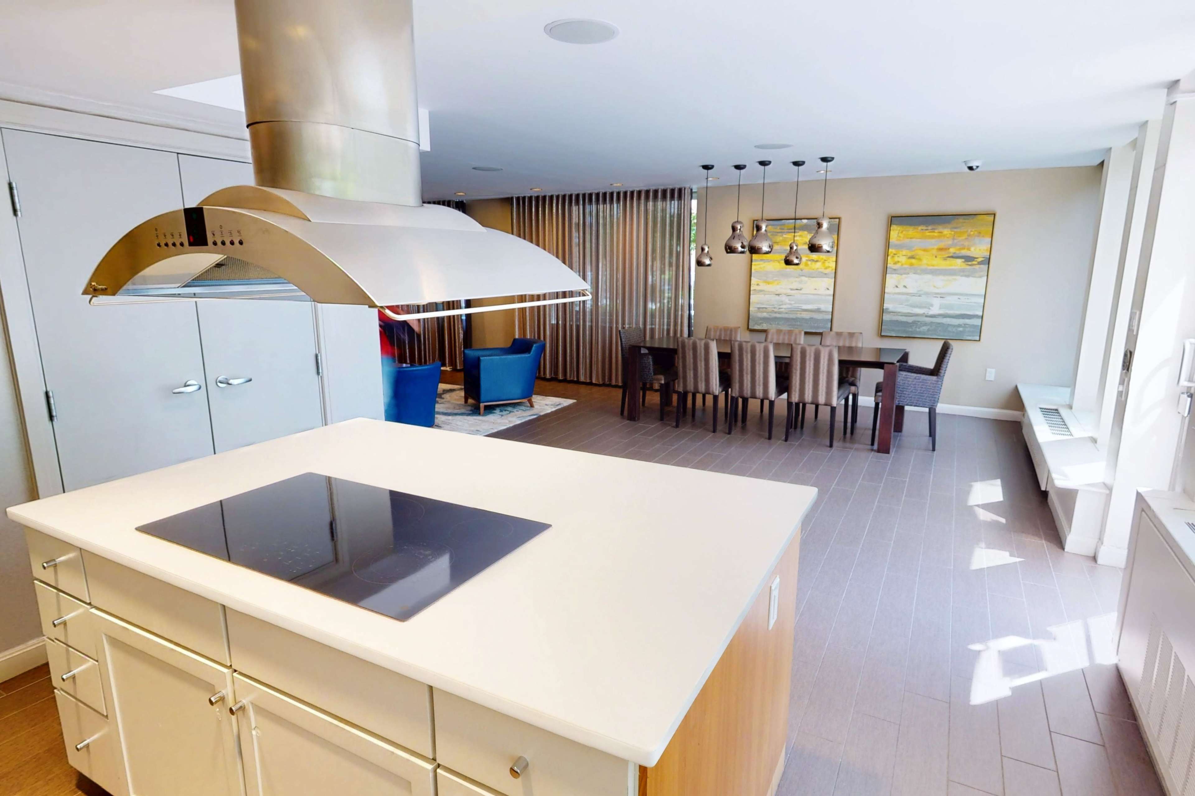 The image shows a modern kitchen with a central island featuring a stovetop, overlooking a dining area furnished with a wooden table and chairs, illuminated by decorative lights.