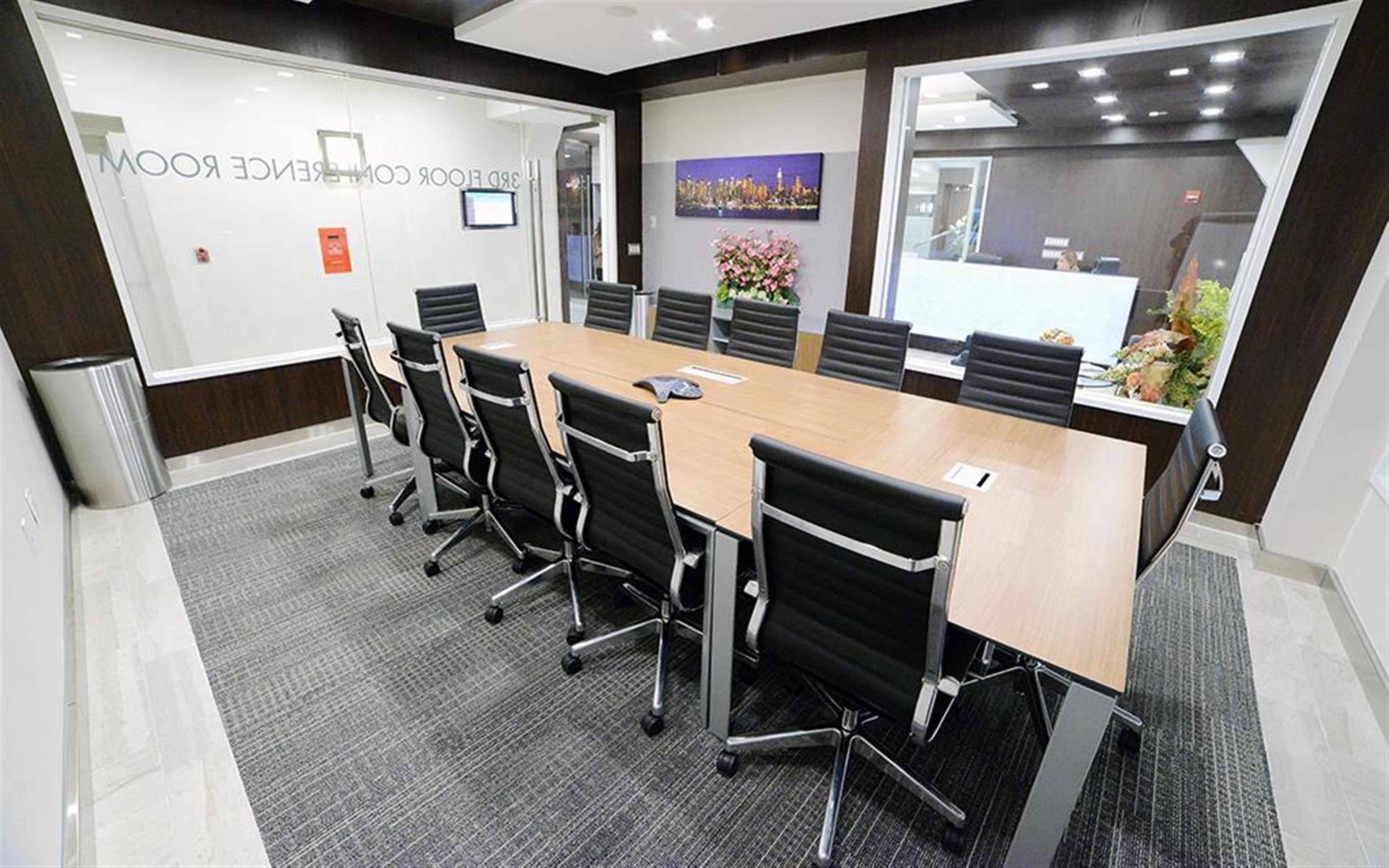 A large conference room features a long wooden table surrounded by black leather chairs, with a glass wall displaying a floral arrangement and a digital screen.