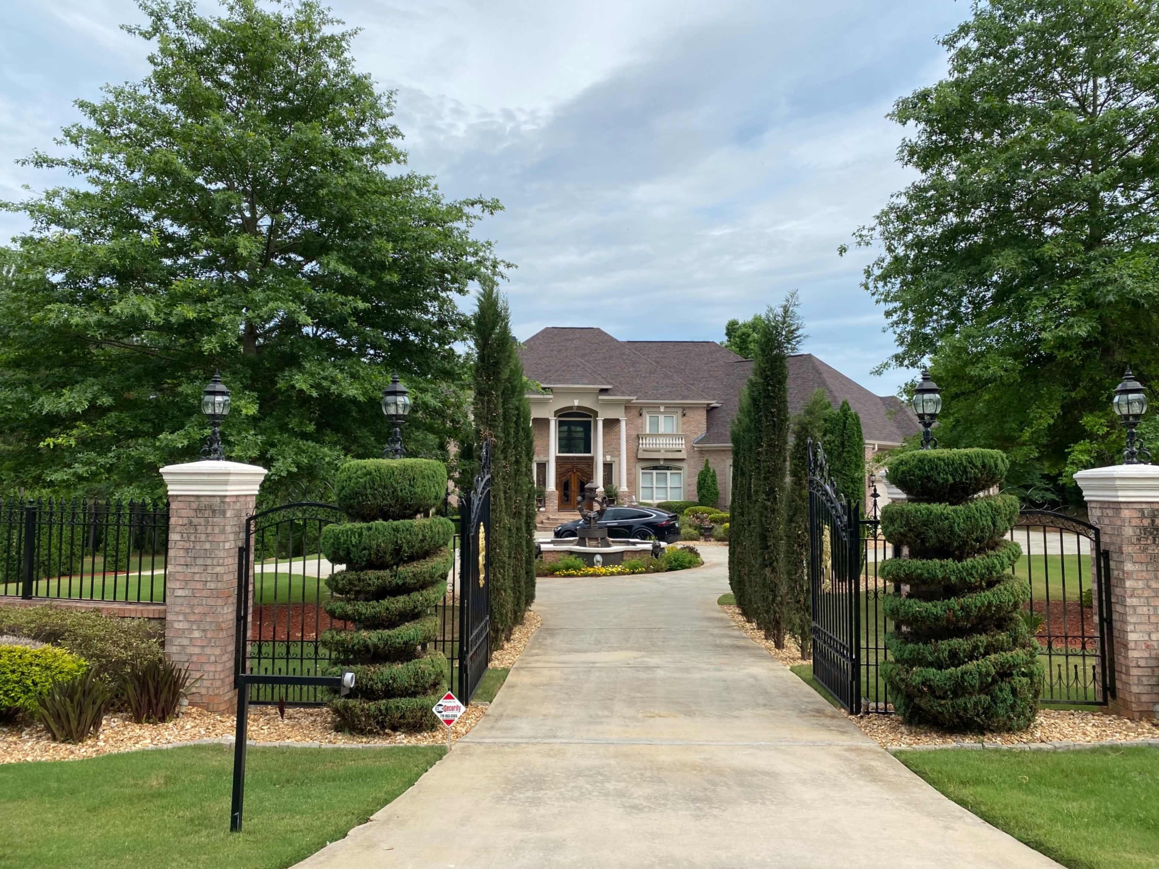 A long driveway leads to a large brick house surrounded by neatly trimmed trees and manicured landscaping.