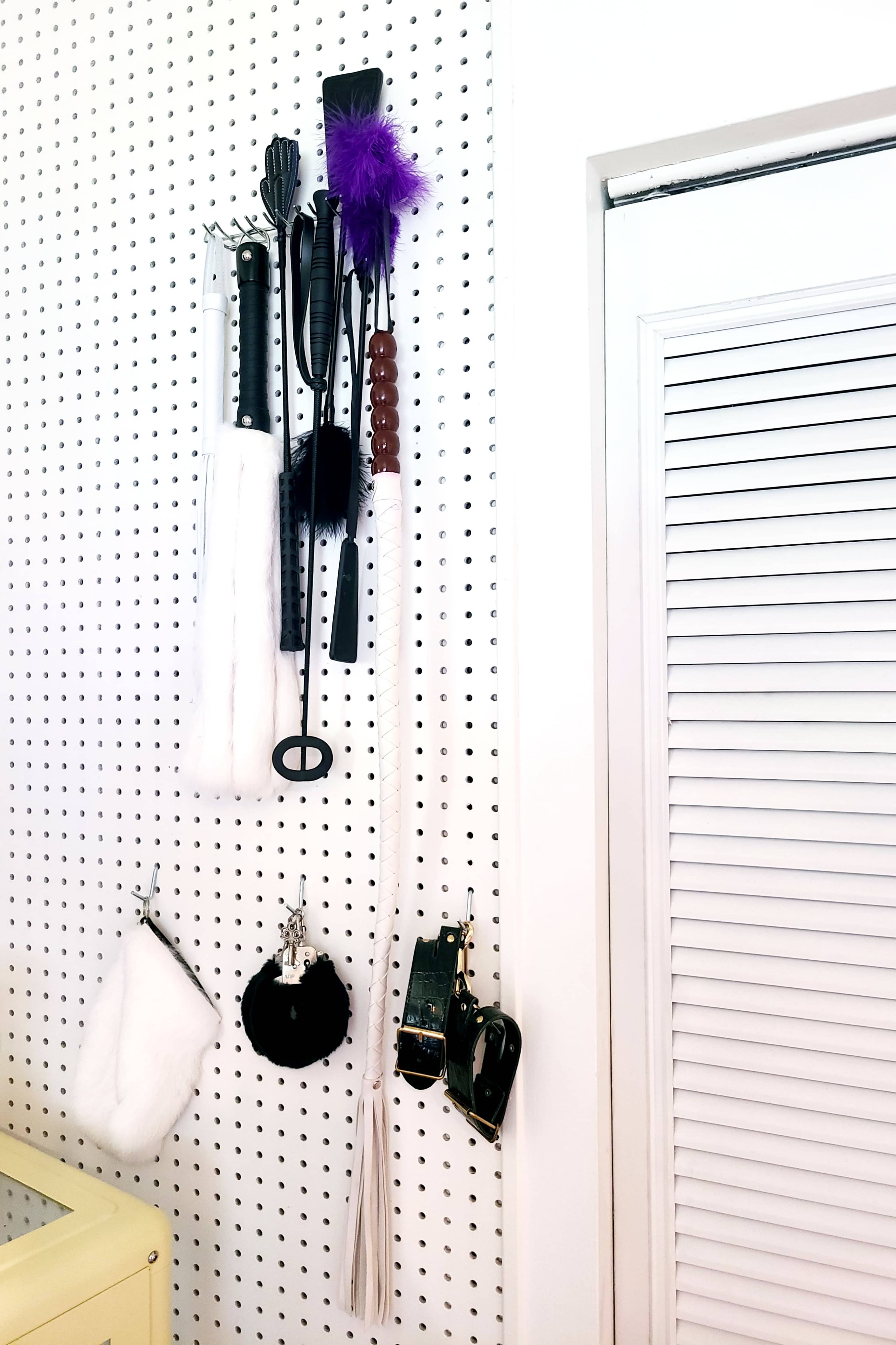 A wall-mounted pegboard with various tools and accessories, including whips, cuffs, and a feather, displayed on hooks.