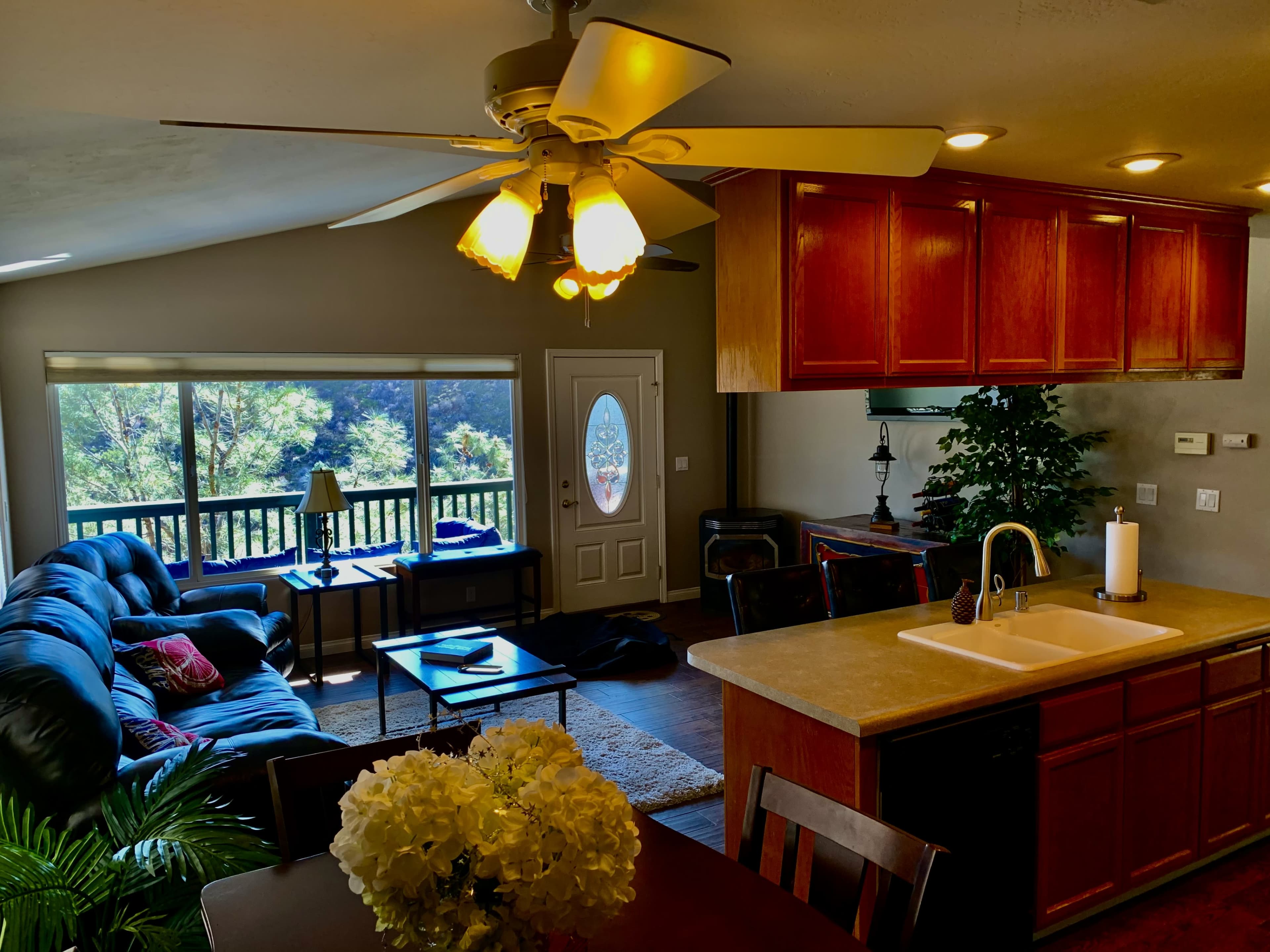 A cozy living room with a large window, a dark leather sofa, a wooden dining table, and a ceiling fan overhead.