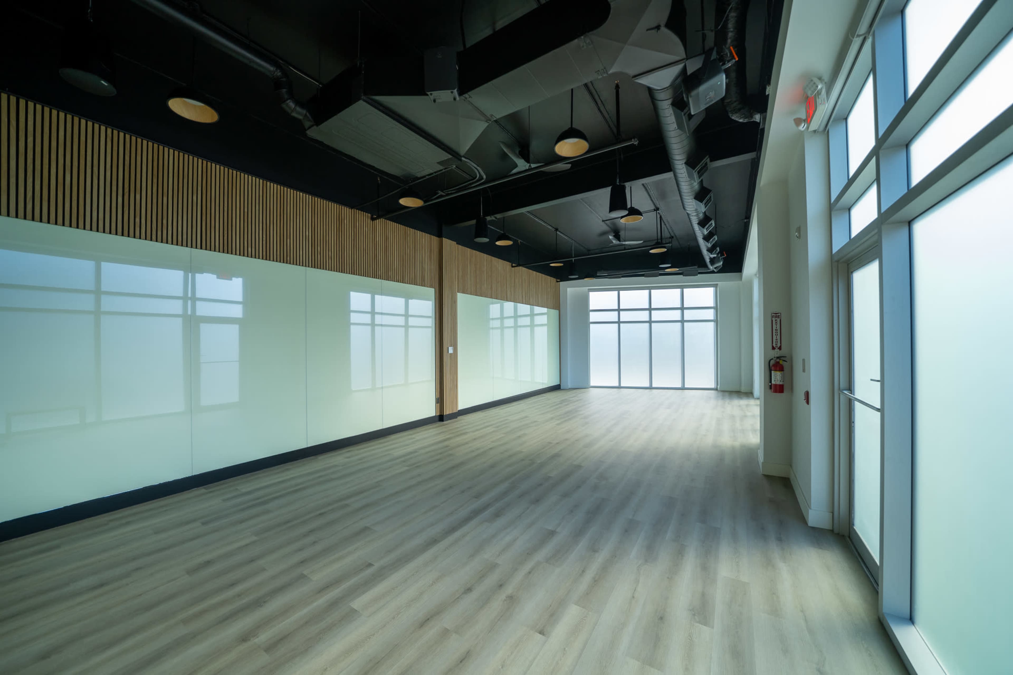 The image shows an empty, well-lit interior space with large windows and wooden flooring.