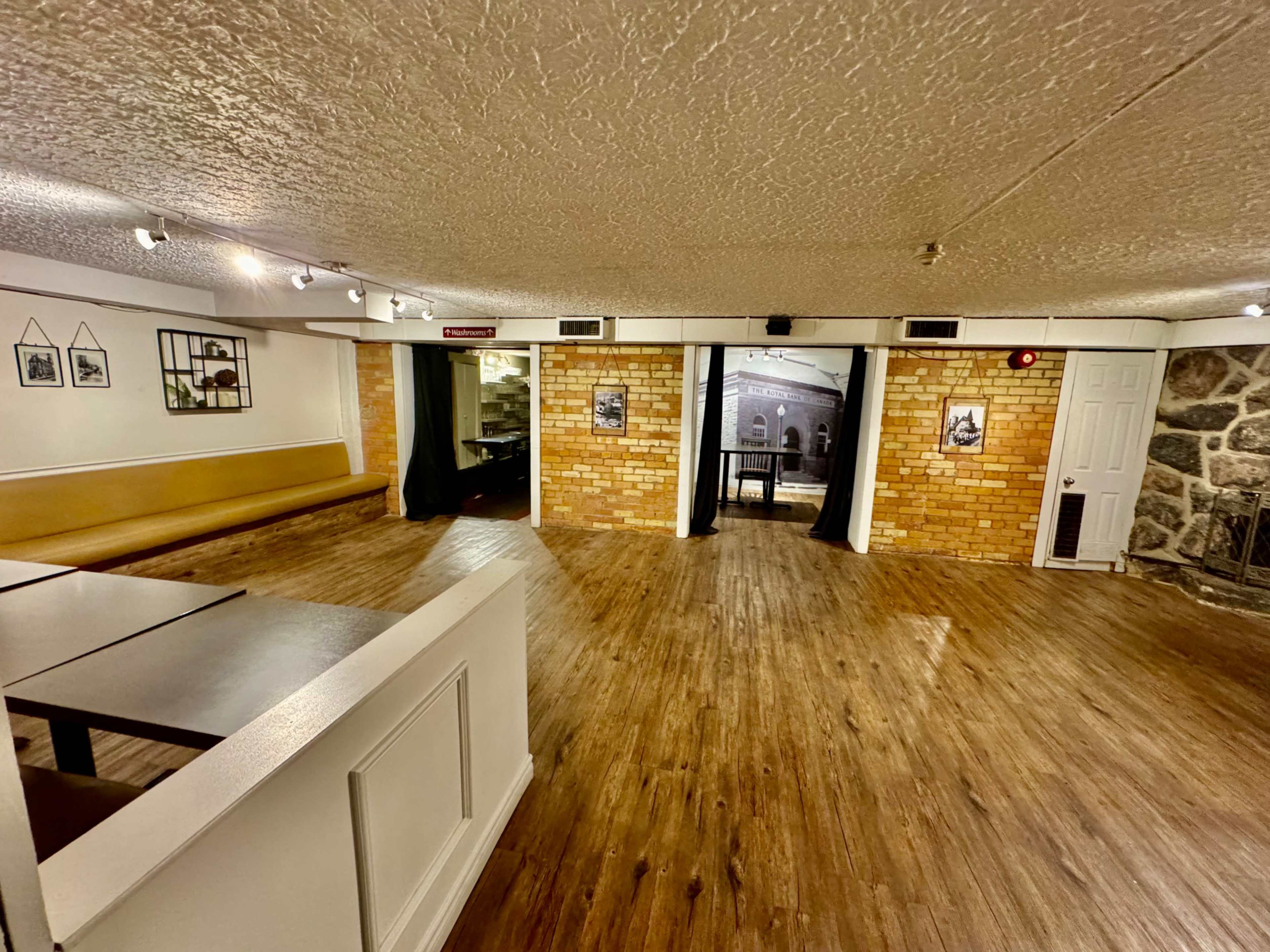 A spacious interior room with wooden flooring, brick walls, and a seating area on one side, leading to a doorway draped with curtains.
