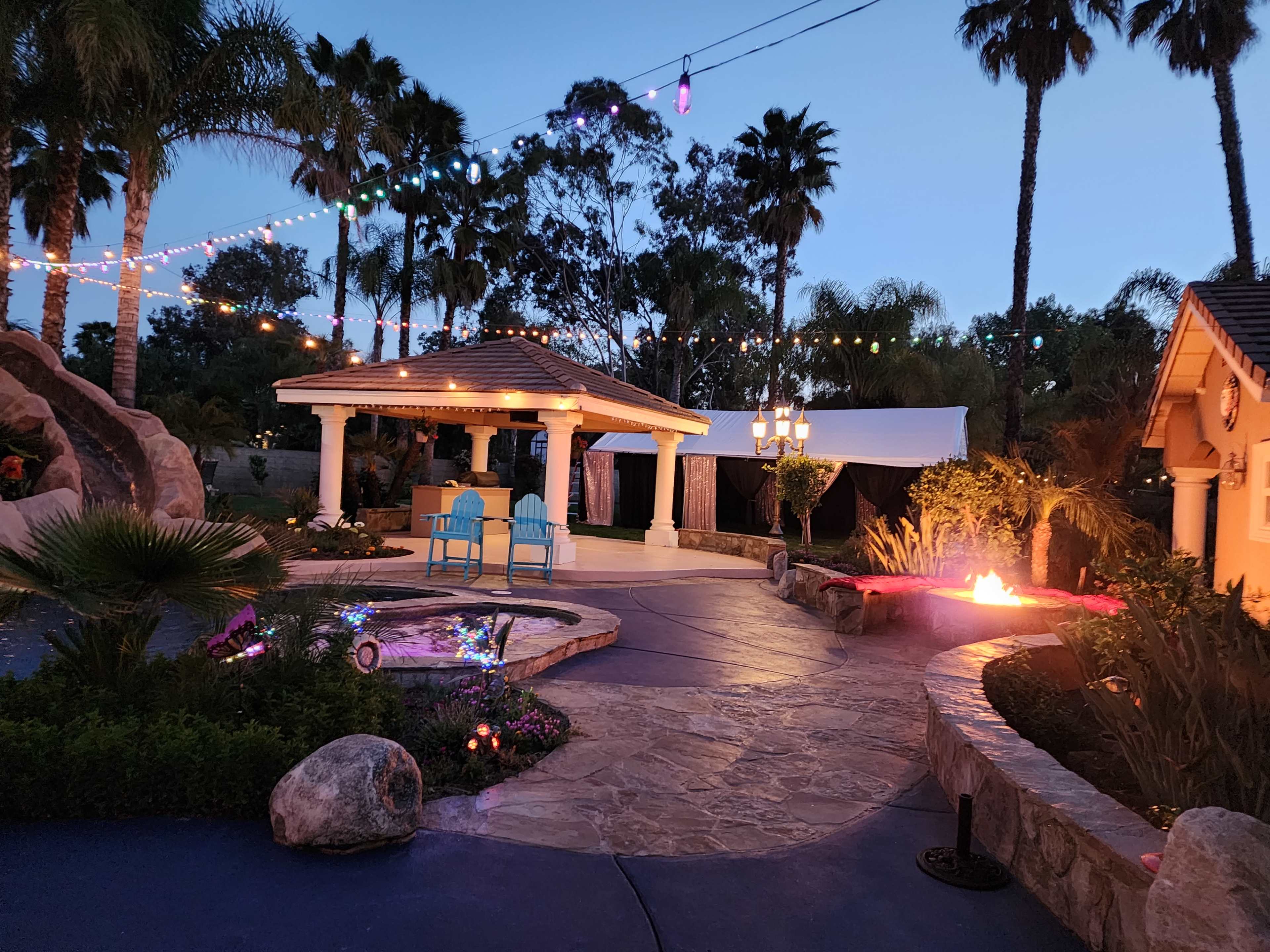 A landscaped outdoor area featuring a gazebo, two blue chairs, and decorative lighting amidst palm trees and a fire pit.