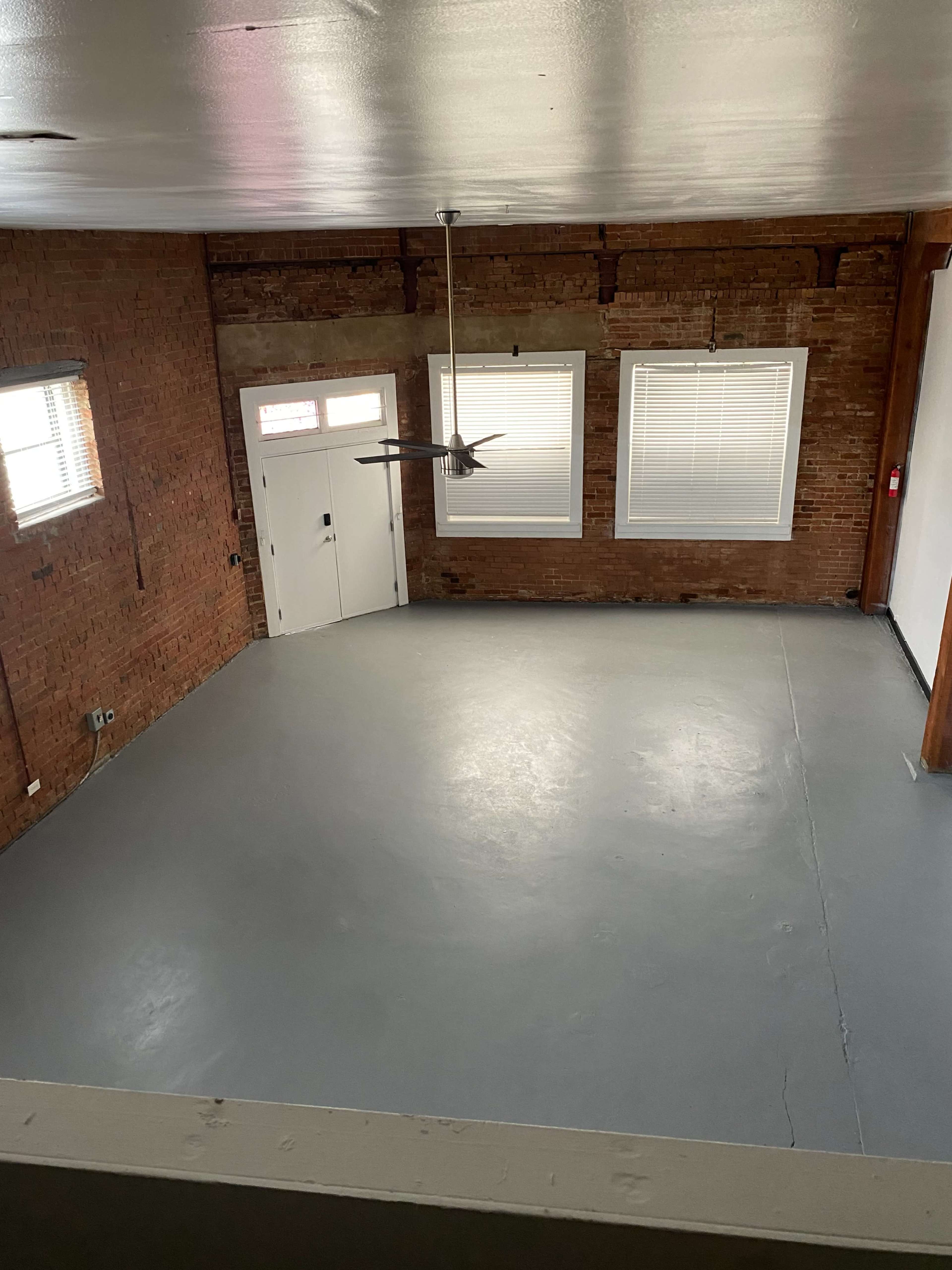 A spacious room with exposed brick walls, a concrete floor, and large windows allowing natural light.