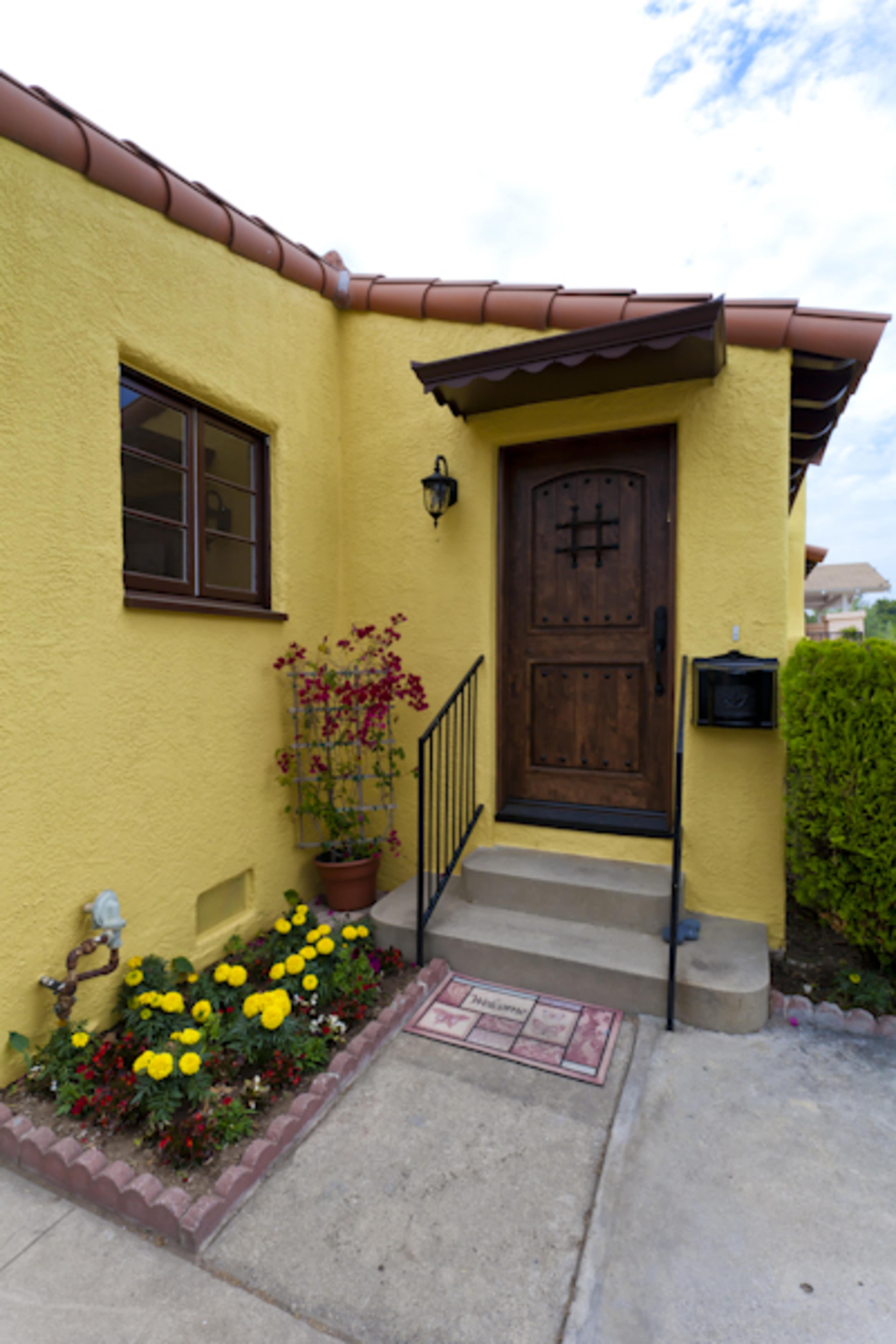The image shows a yellow stucco house with a wooden front door, a small set of stairs, and a flower bed filled with blooming plants beside the entrance.