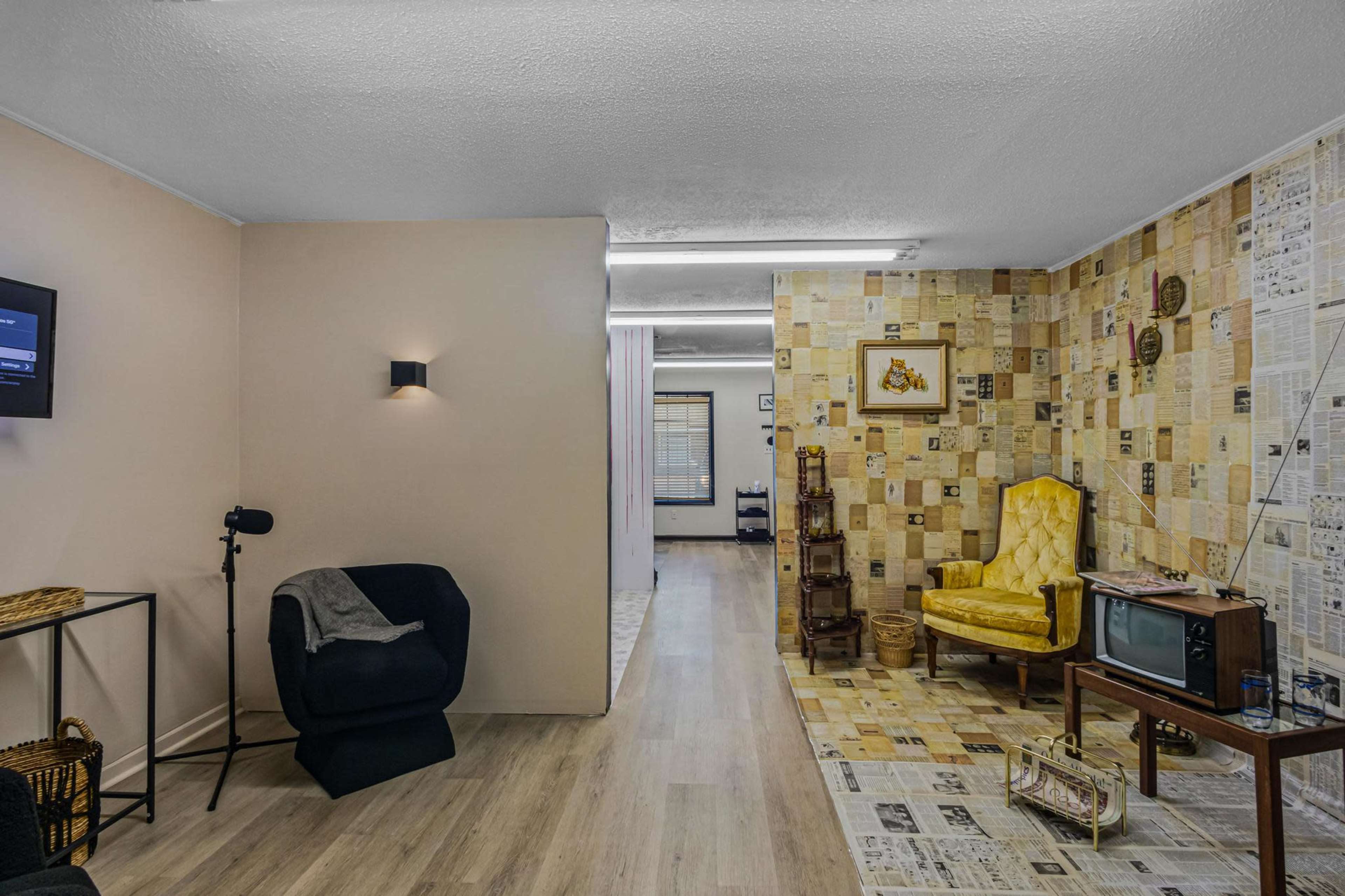 A room with vintage decor, featuring a yellow armchair, a TV on a stand, and walls partially covered with newspaper-like wallpaper.