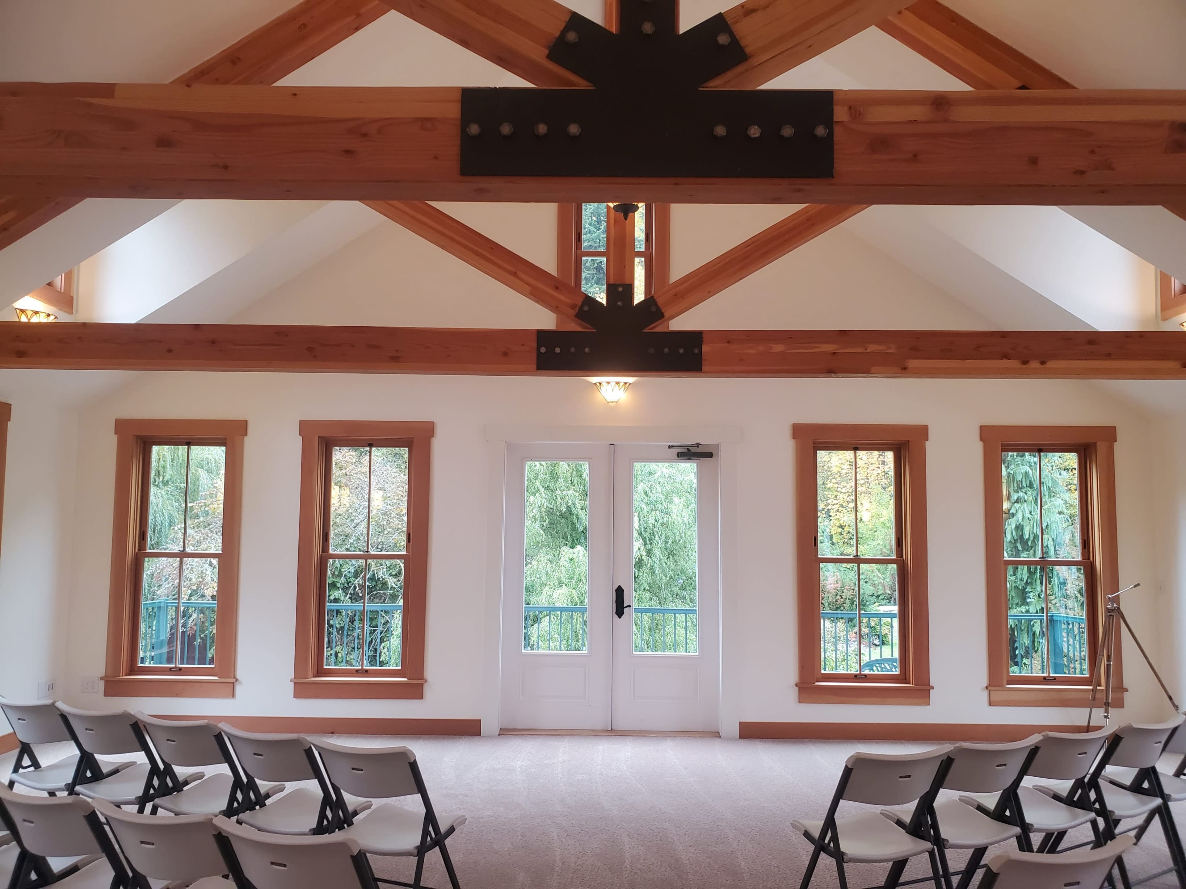 The image shows a spacious room with wooden beams, large windows, and rows of folding chairs facing a pair of doors.