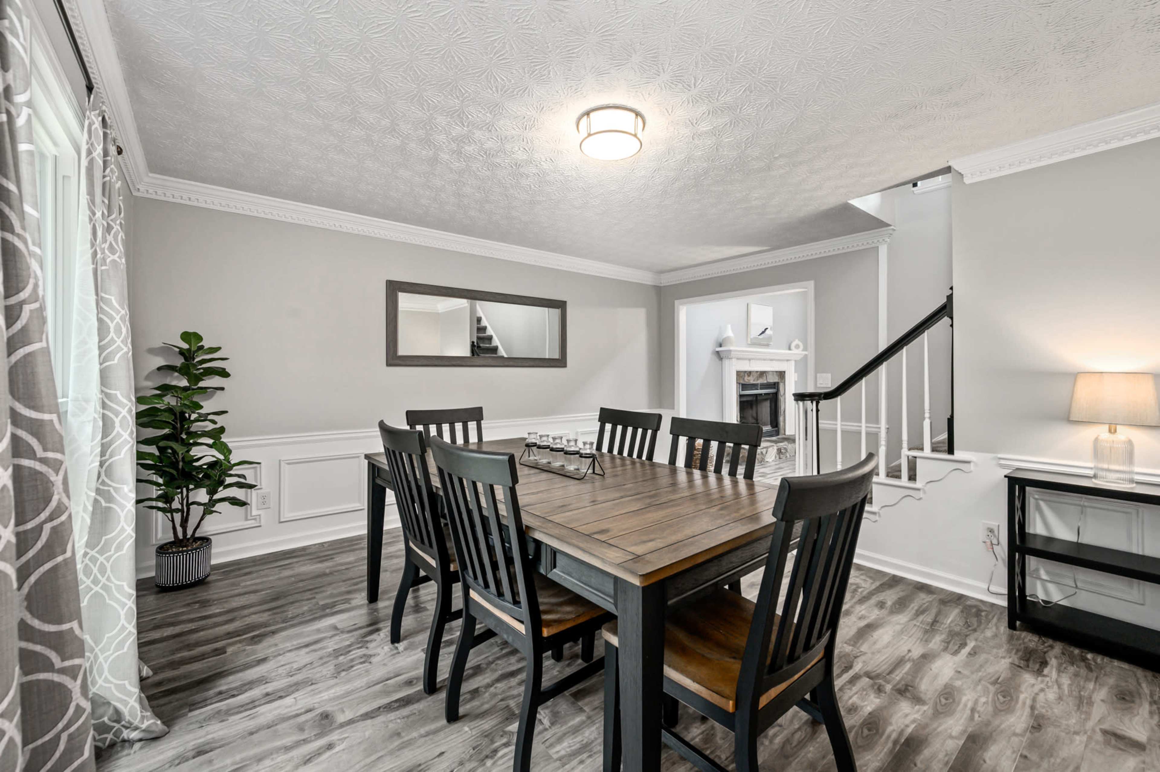 A dining room featuring a wooden table surrounded by black chairs, with gray walls and decorative molding, alongside a staircase and a fireplace in the background.