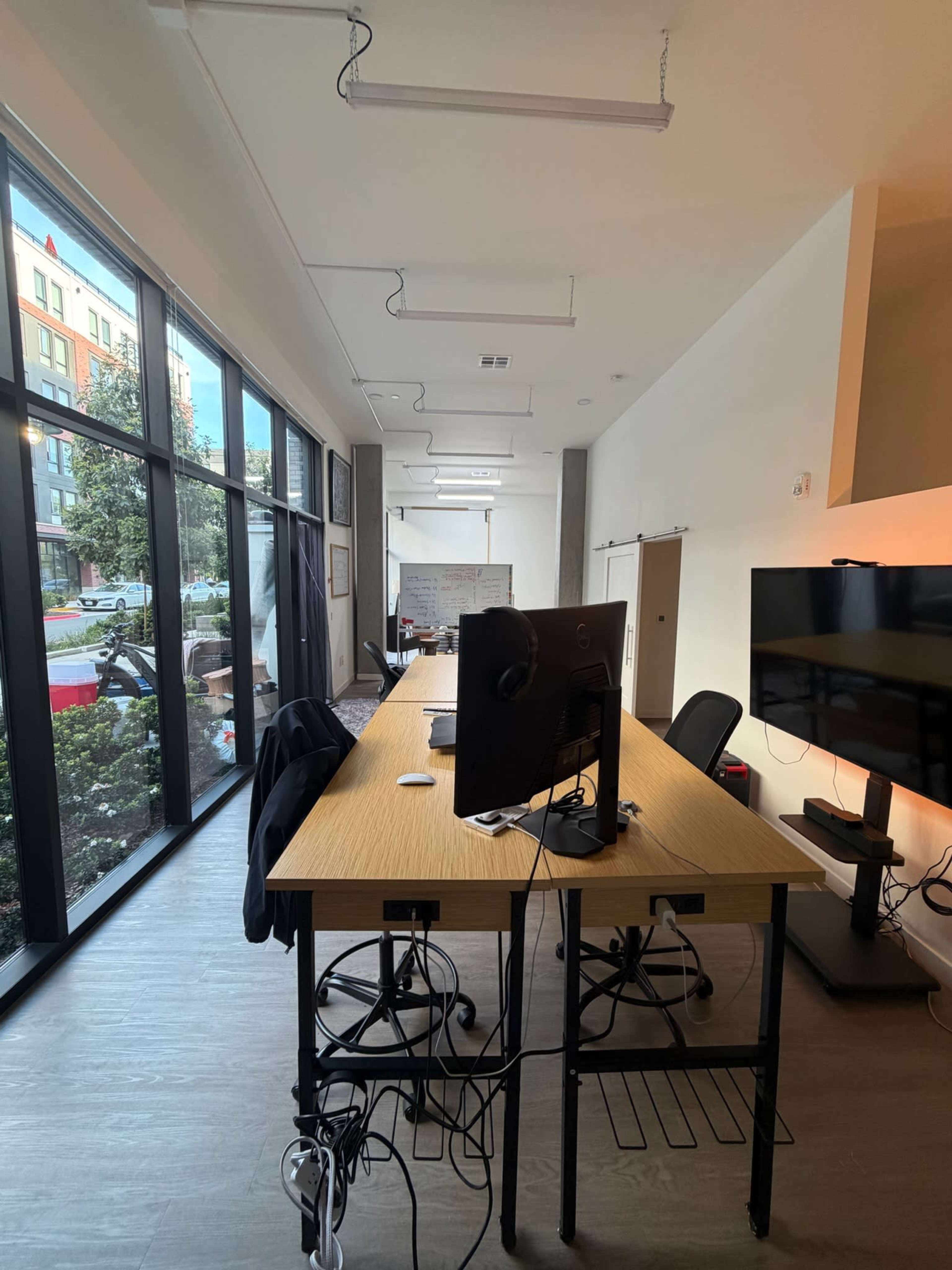 The image shows a modern office space with a long table, several chairs, and large windows allowing natural light to enter.