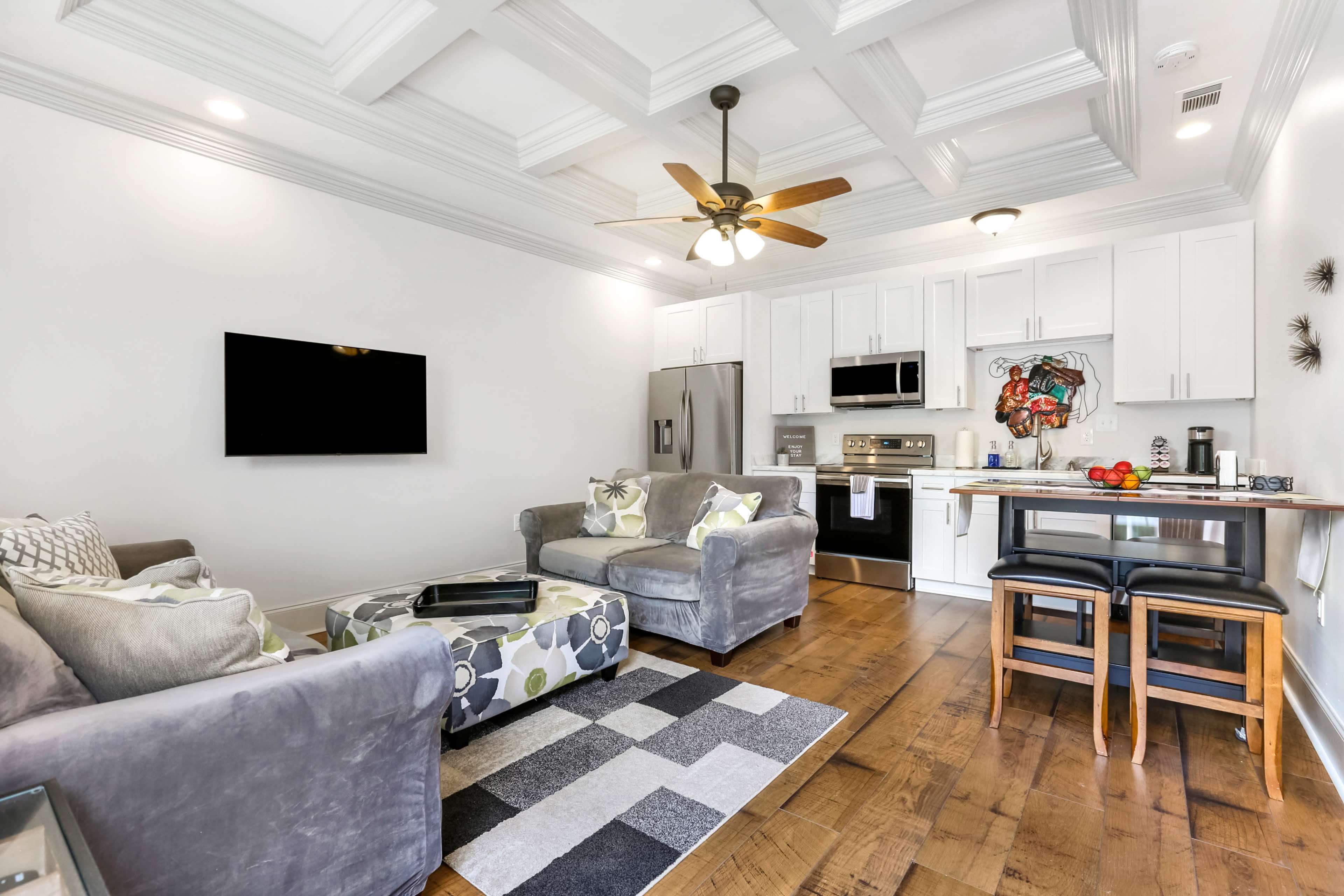 The image shows a modern living area with a gray sectional sofa, a small dining table with black stools, and a kitchen featuring white cabinets and stainless steel appliances.