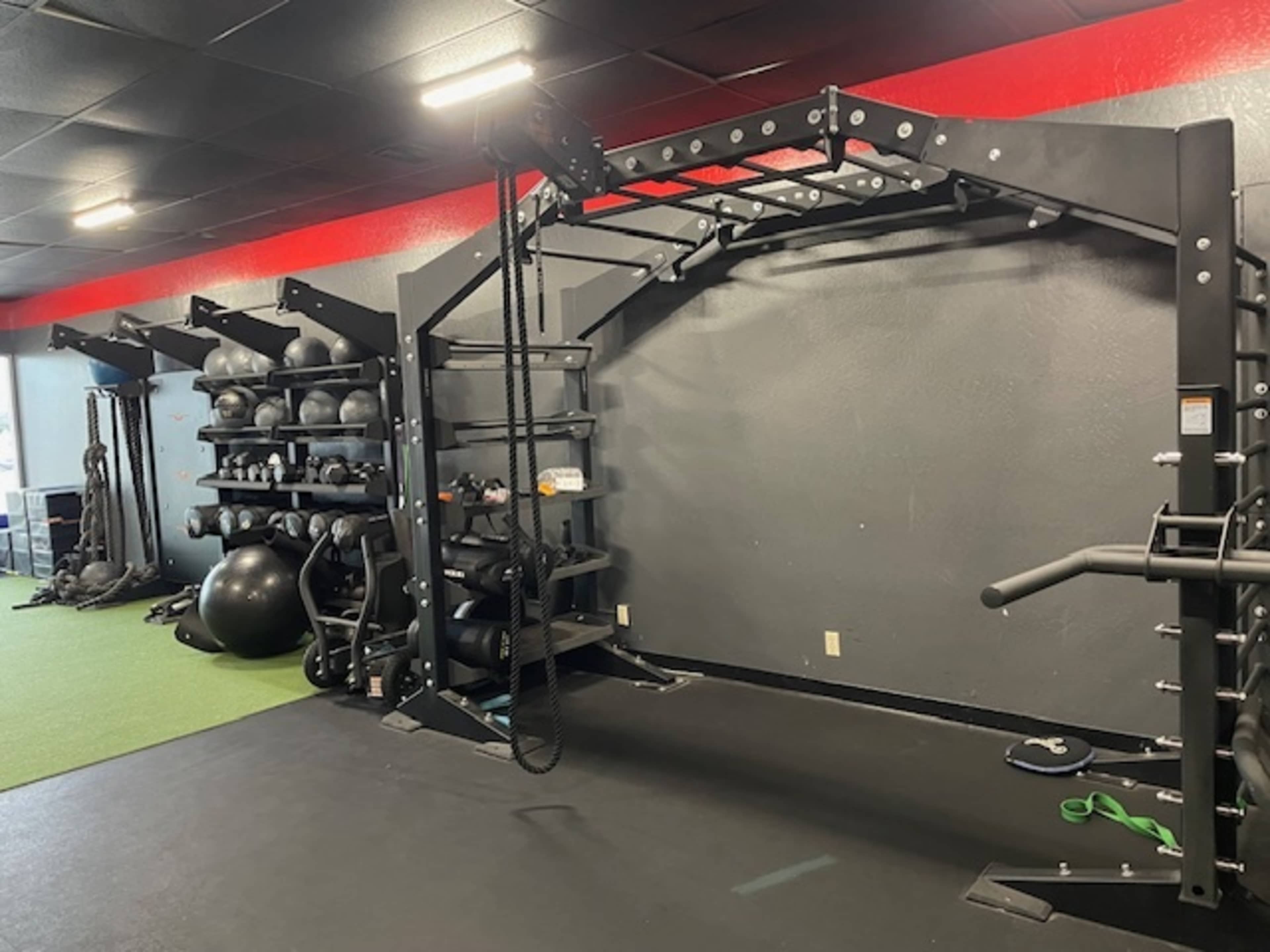 A gym interior with a black multi-functional rig, storage for fitness equipment on the wall, and various exercise tools on the floor.