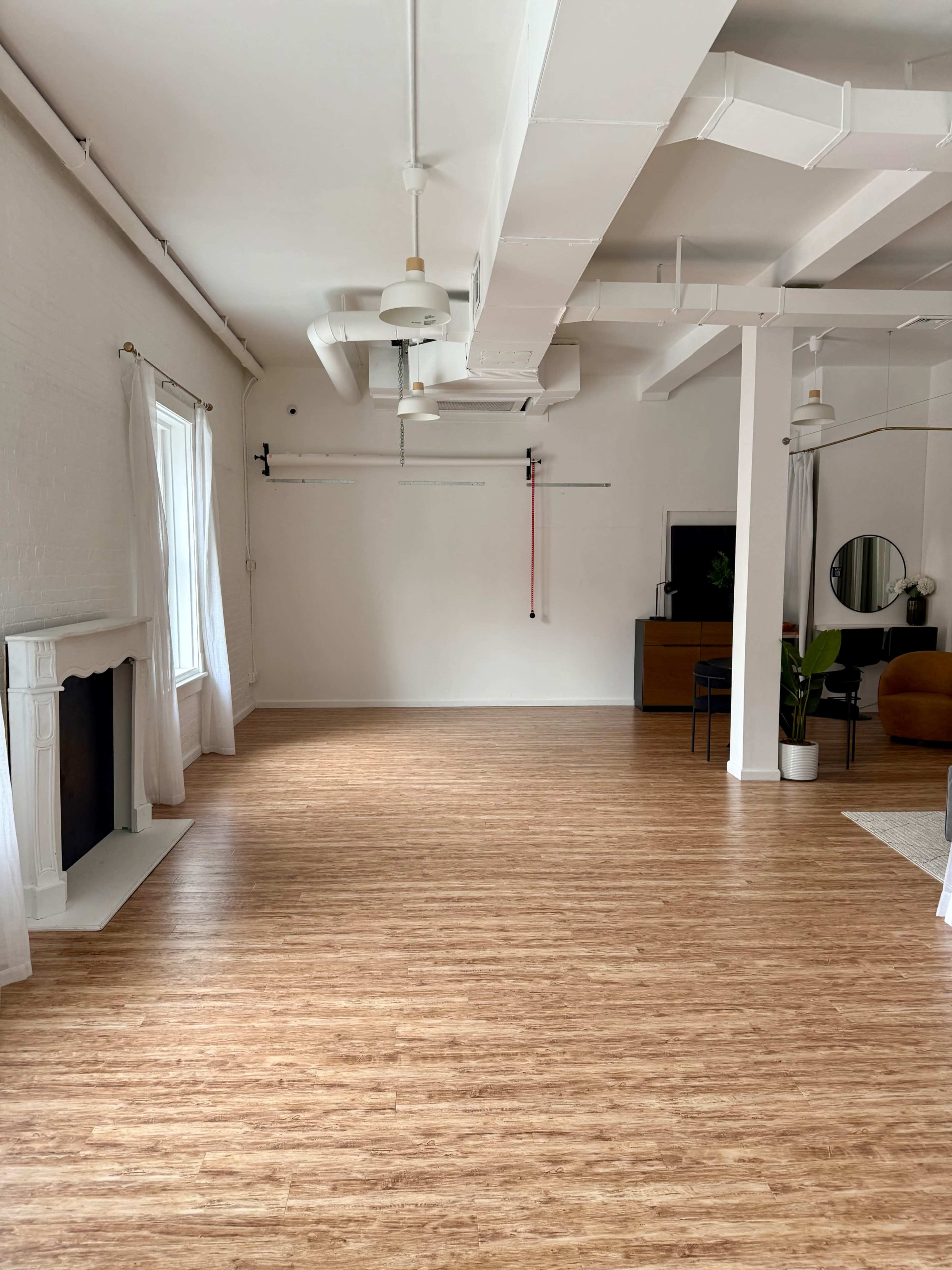 An open, spacious studio features wooden flooring, large windows with sheer curtains, and minimal furniture, including a chair and a small table.