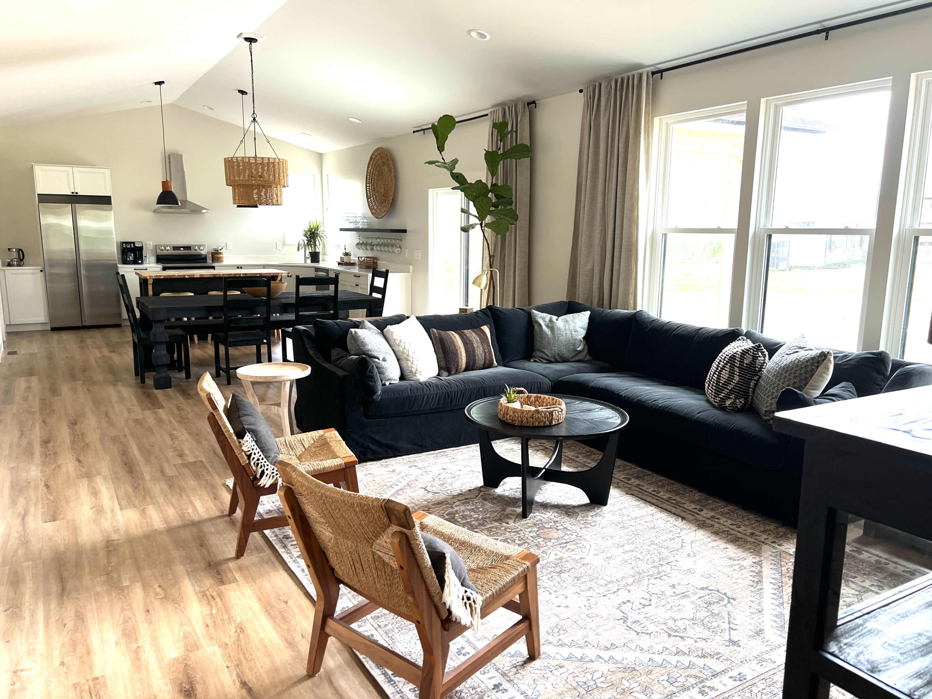 The image shows an open-concept living space featuring a black sectional sofa, a round coffee table, and a dining area with a wooden table and chairs.