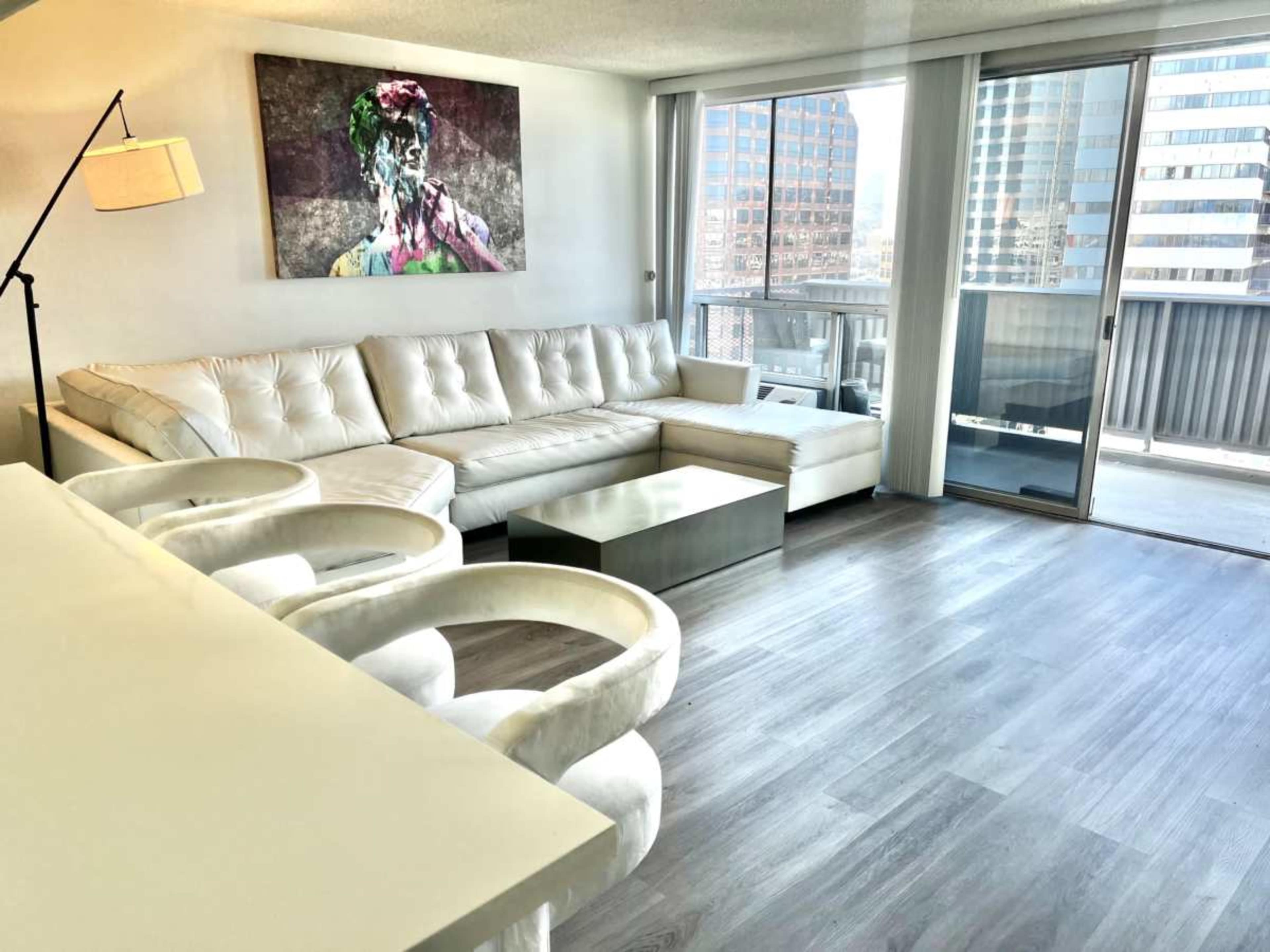 The image shows a modern living room with a white sectional sofa, a glass coffee table, and large windows that offer a view of a city skyline.