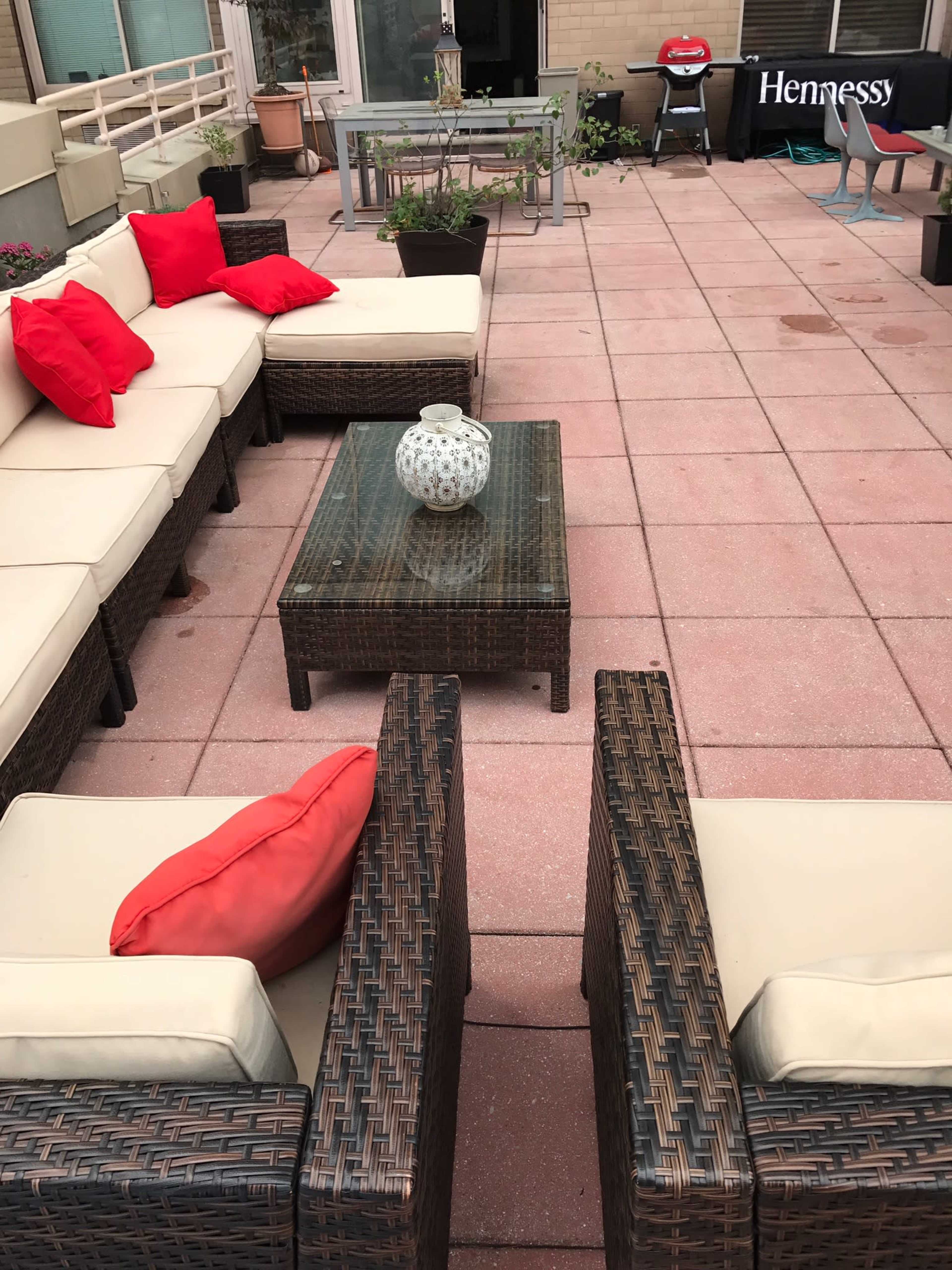 The image shows a patio area with a sectional sofa, a coffee table, and several potted plants, featuring red cushions and a Hennessy advertisement in the background.