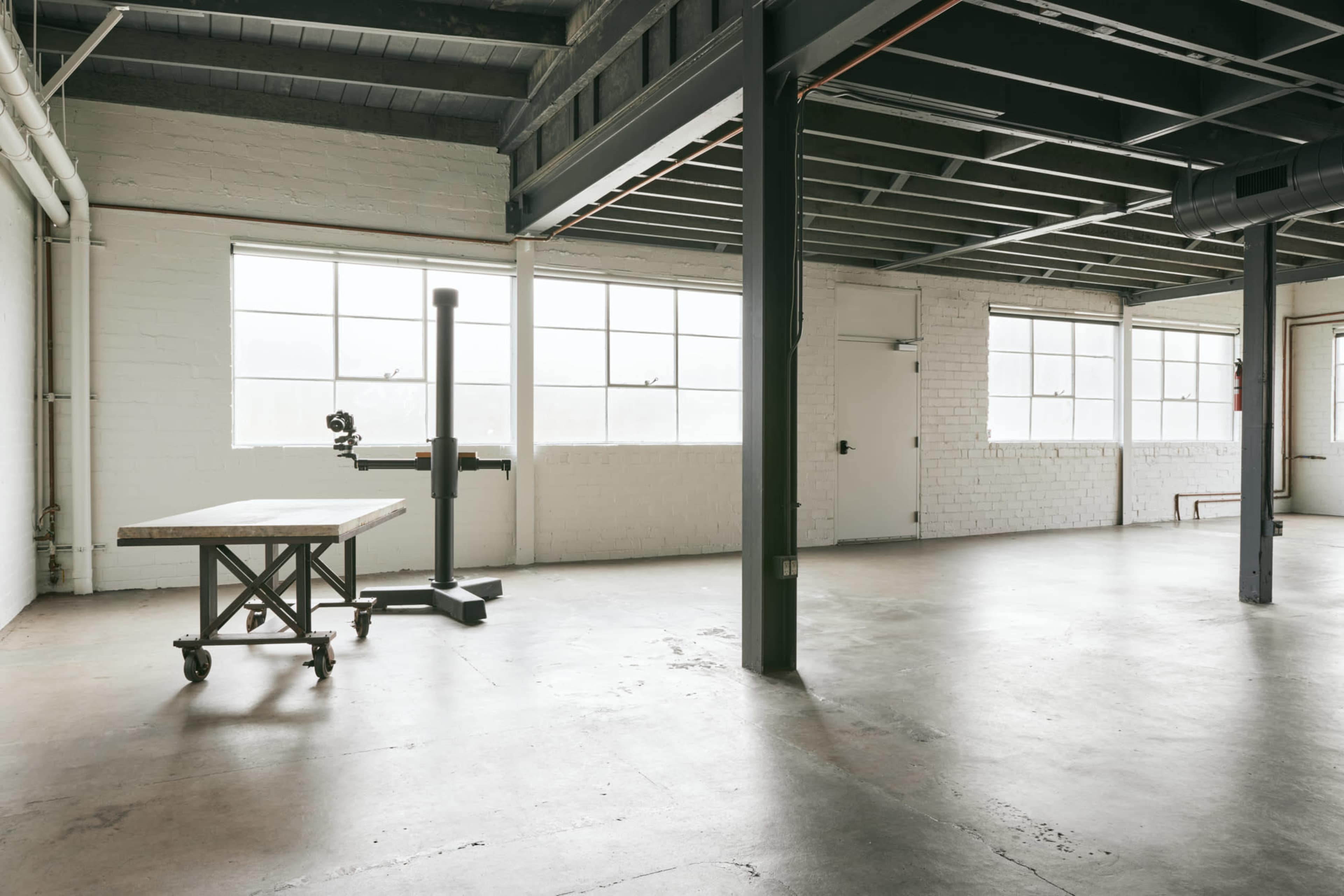An empty industrial space features large windows, a wooden table on wheels, and a vertical stand against a white brick wall.