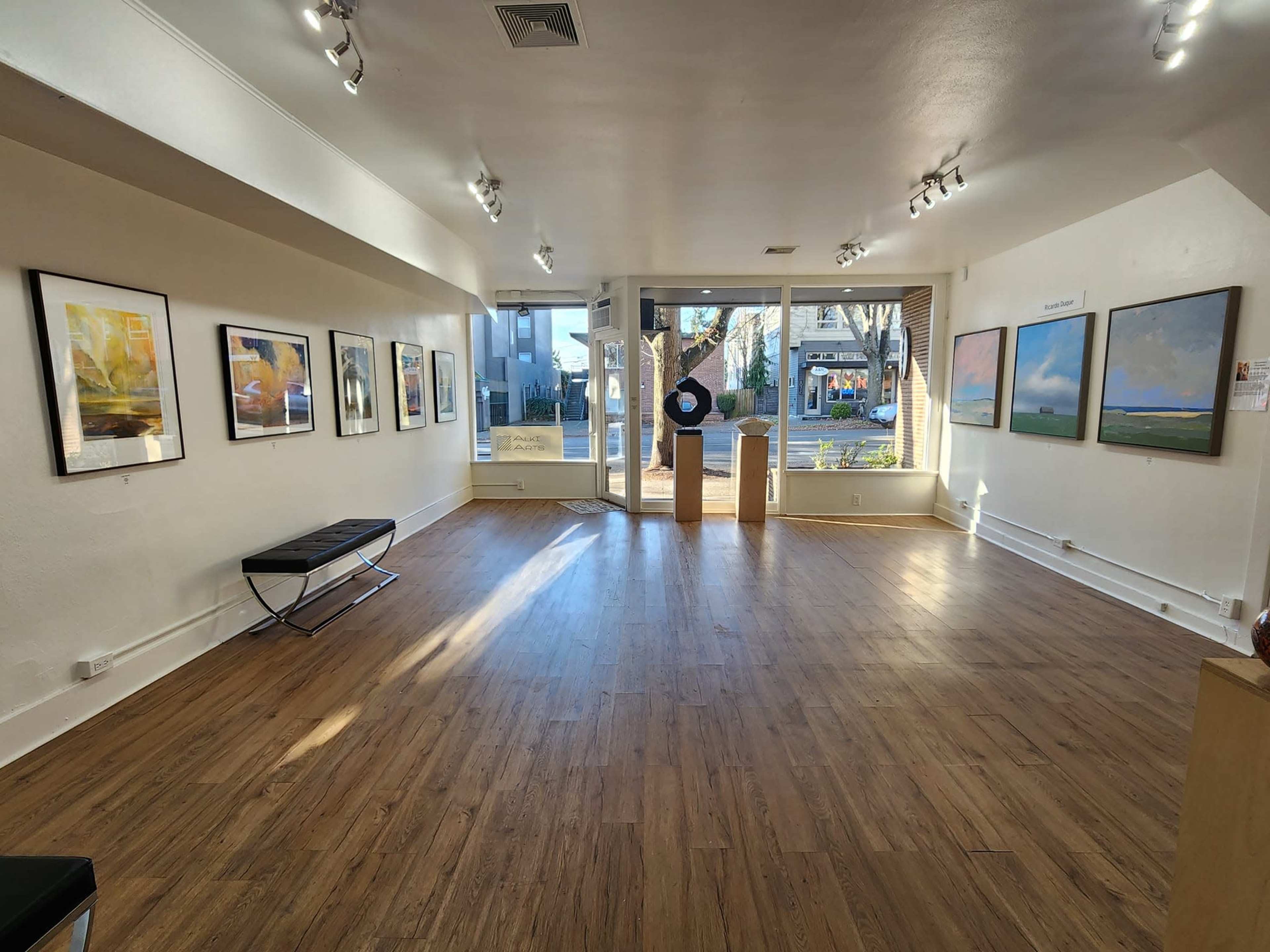An art gallery with wooden flooring and several framed paintings displayed on the walls, leading to a large window at the entrance.
