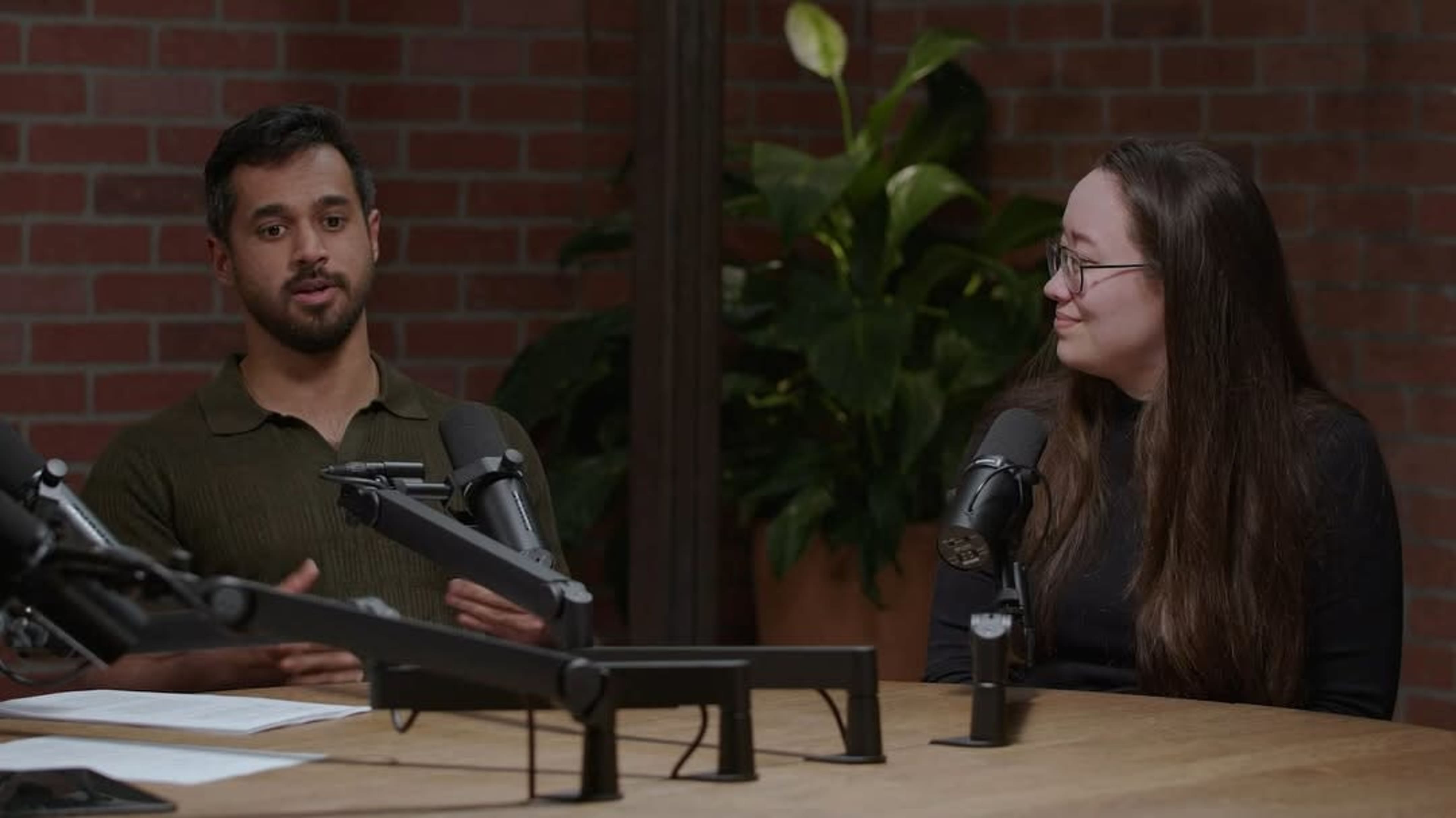 Two individuals are seated at a table with microphones in front of them, engaged in a discussion against a brick wall and partially visible plants.