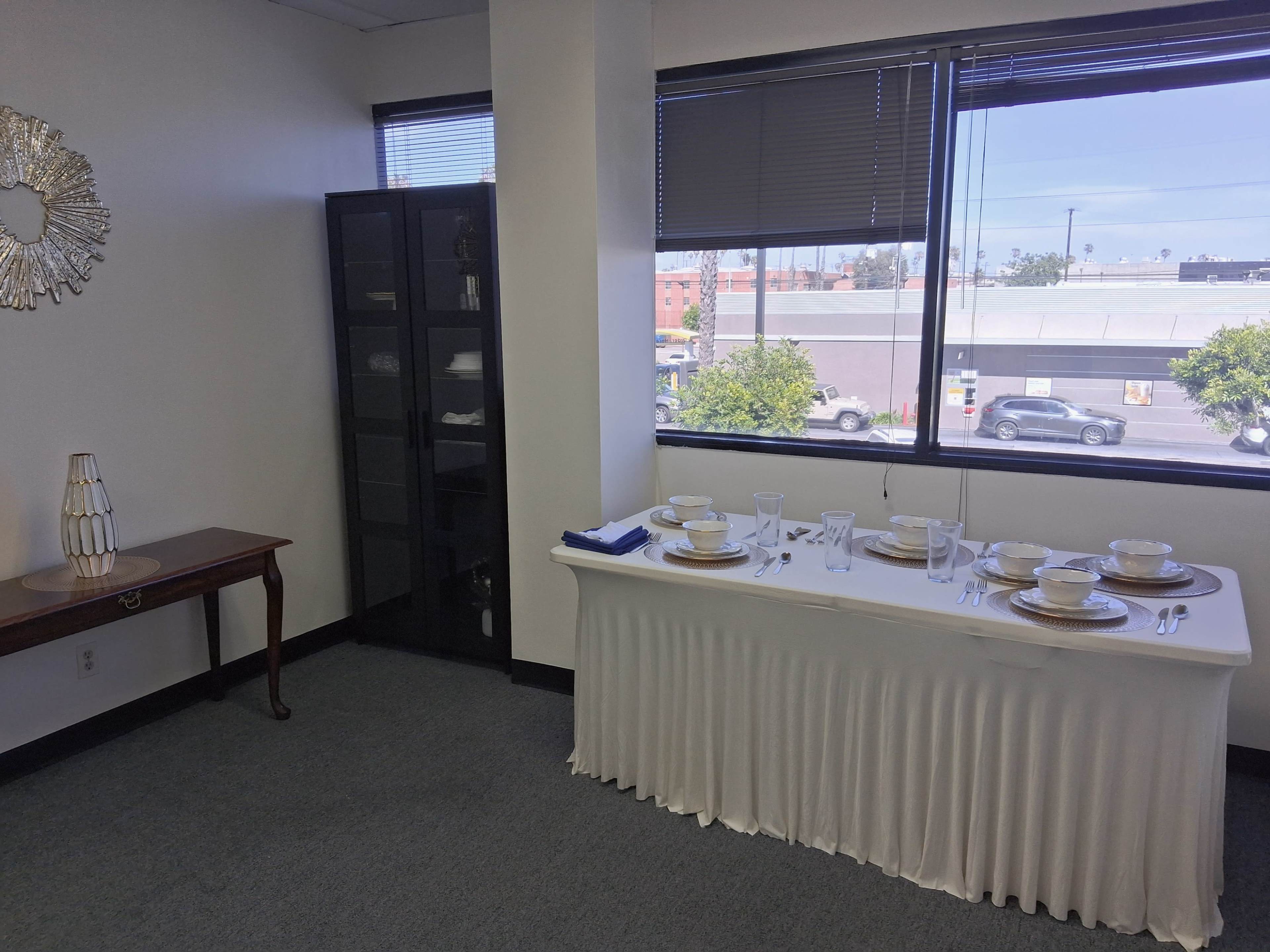 The image shows a neatly set dining table with plates, utensils, and glasses, accompanied by a decorative side table and a display cabinet in an office-like space.
