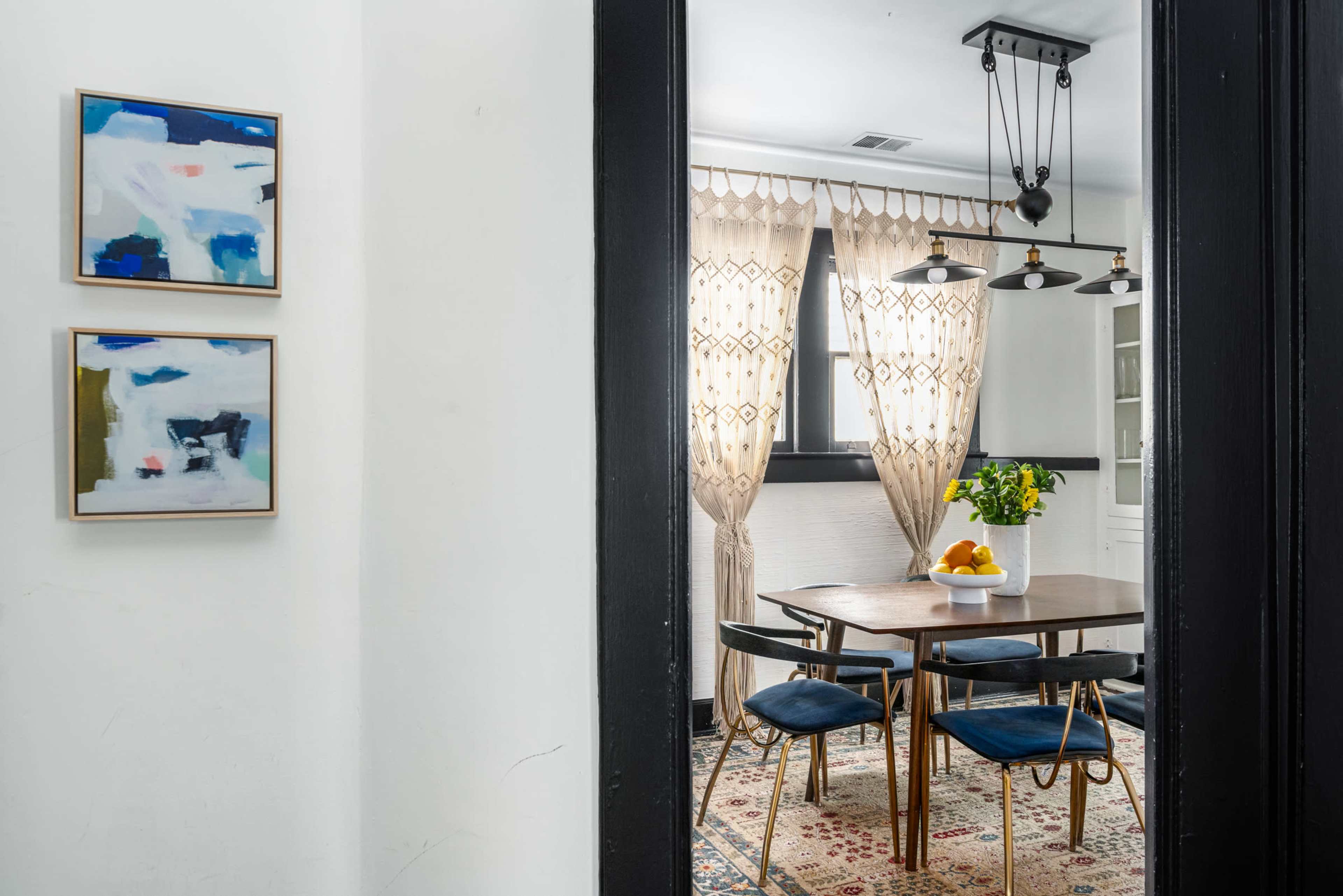A dining area is visible through a doorway, featuring a wooden table, upholstered chairs, and curtains that frame a window.