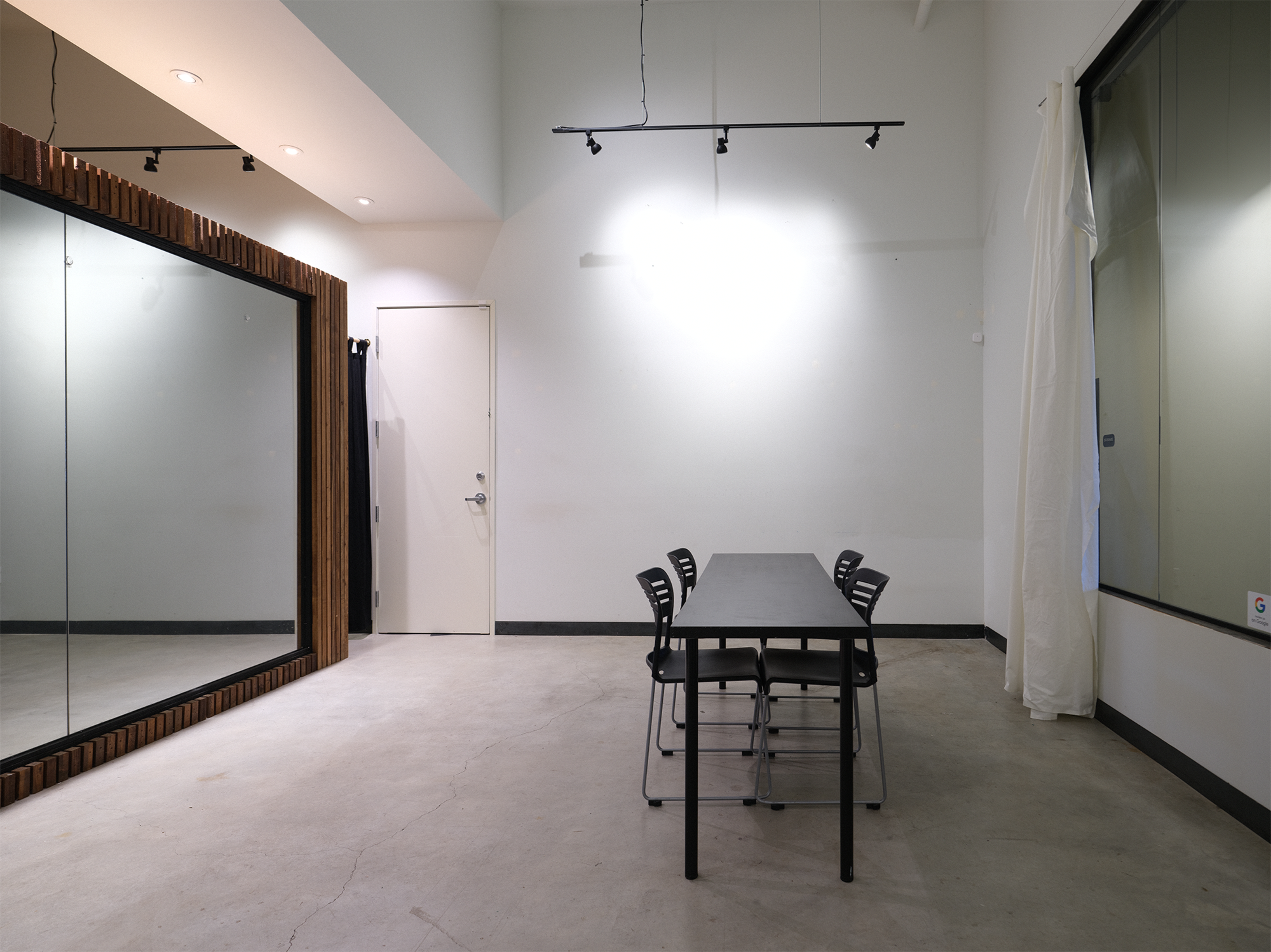A minimalist room features a large mirror, a white wall, and a table surrounded by four black chairs.