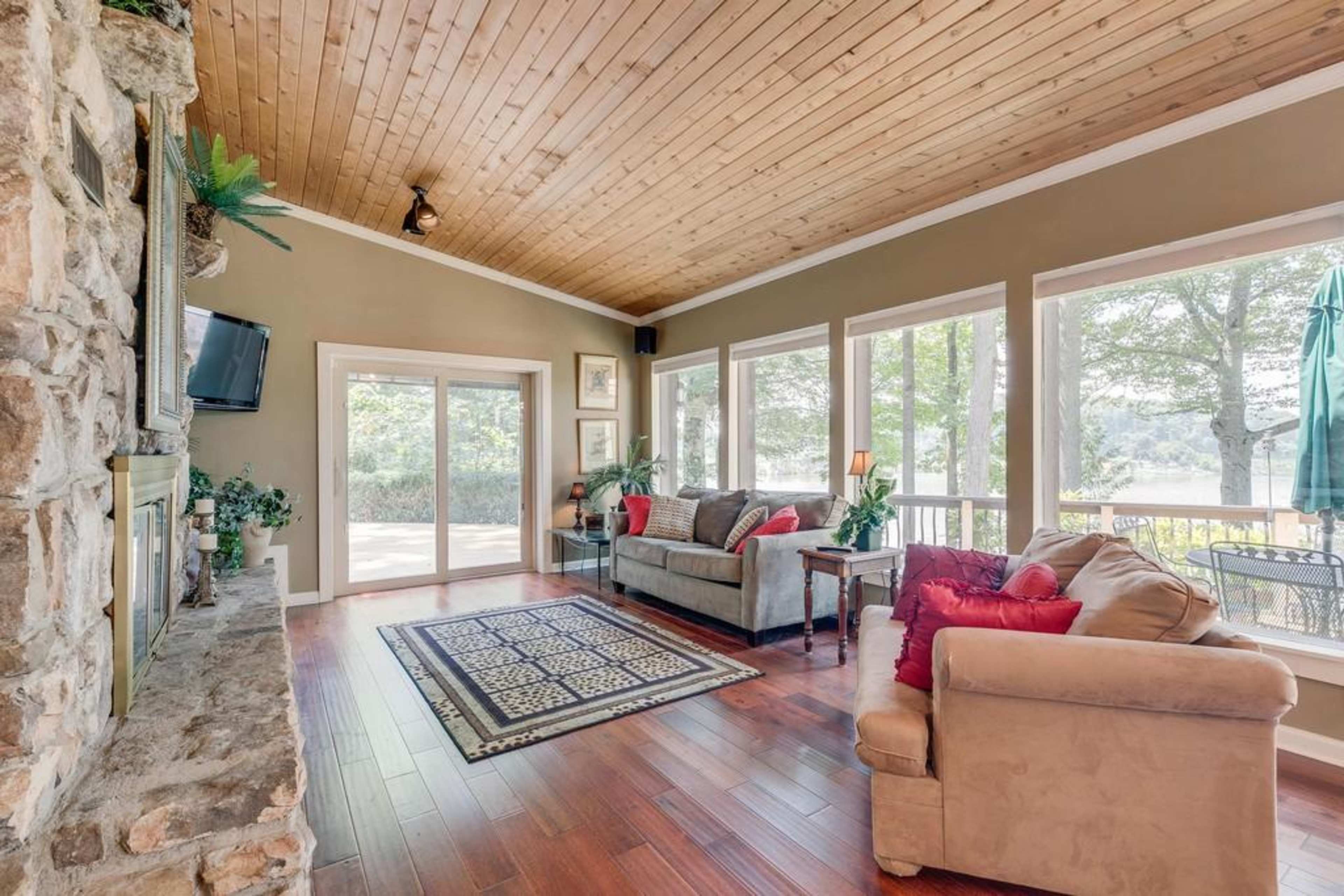 A living room with wooden ceilings, large windows providing a view of the outdoors, and a stone fireplace.