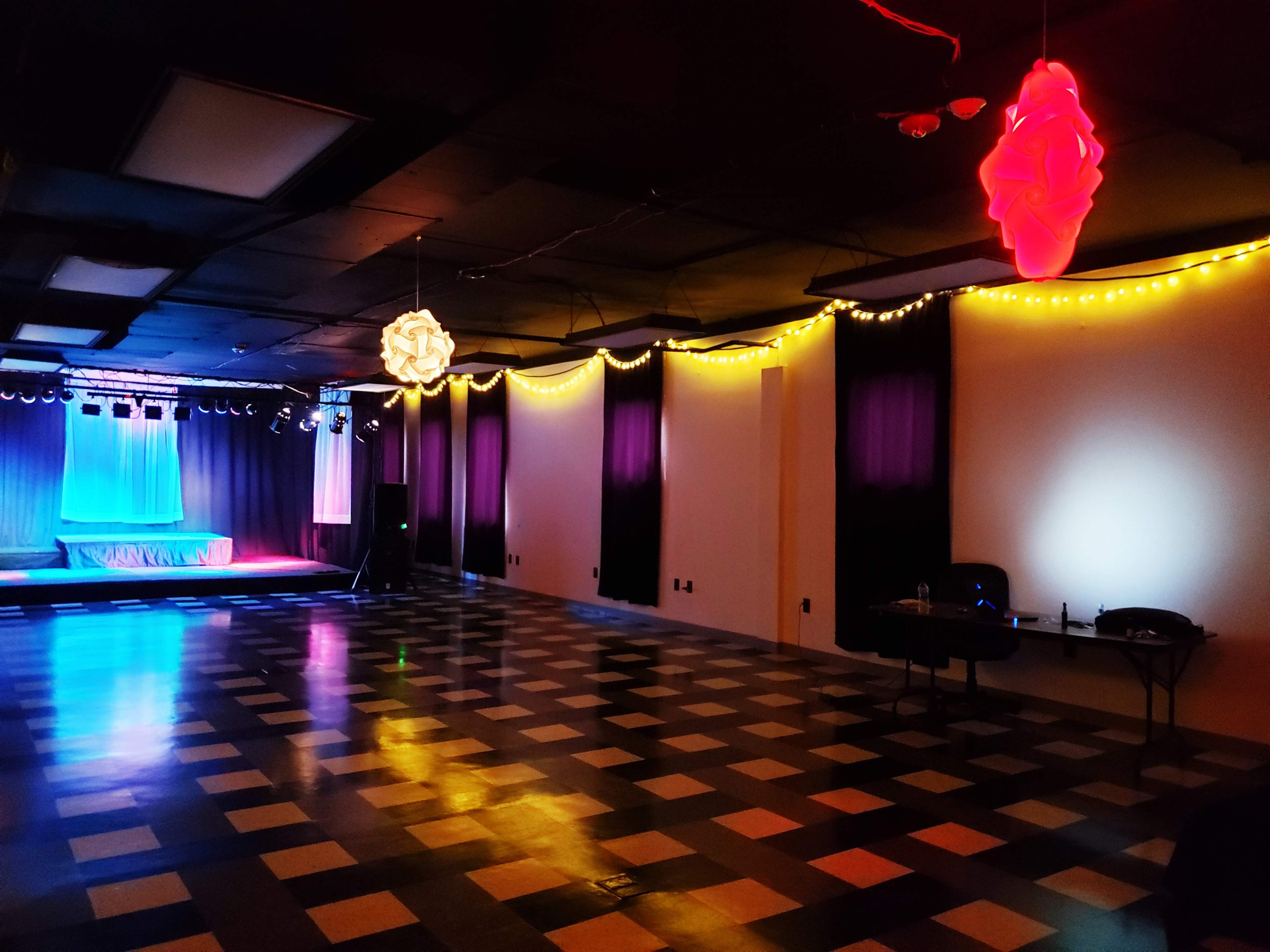 An empty event space with a stage in the background, decorated with colorful lighting and checkered flooring.