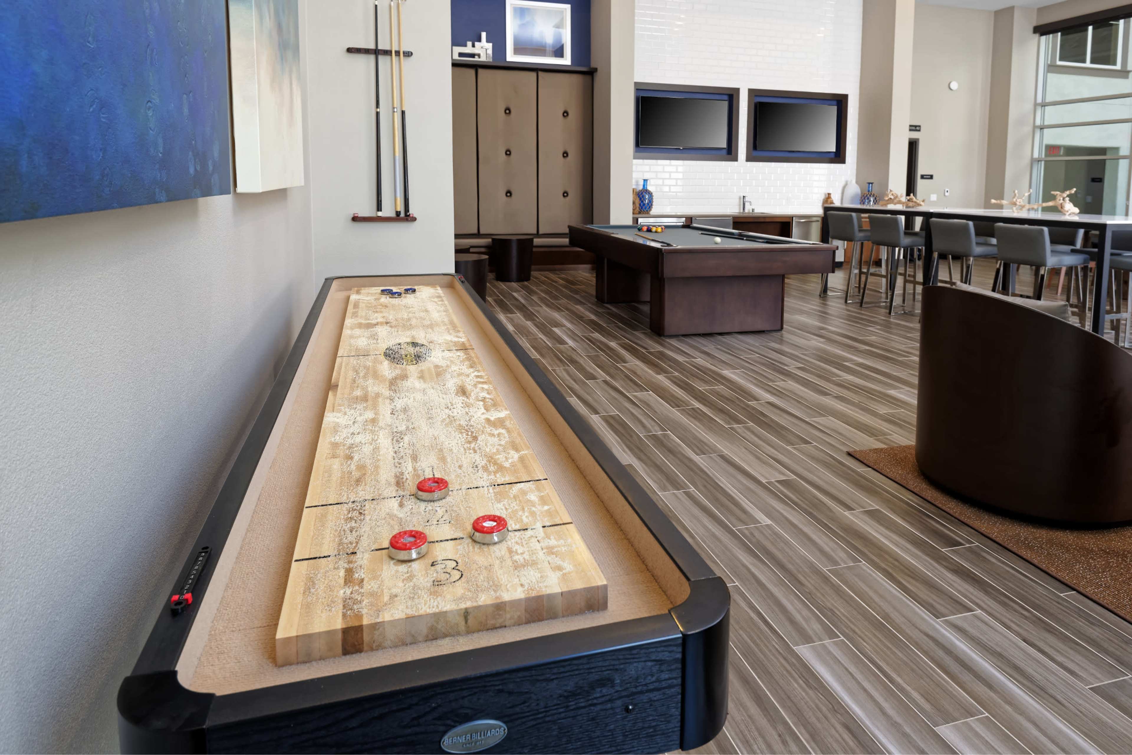 A modern recreational room featuring a shuffleboard table, two pool tables, and a dining area with high-backed chairs.