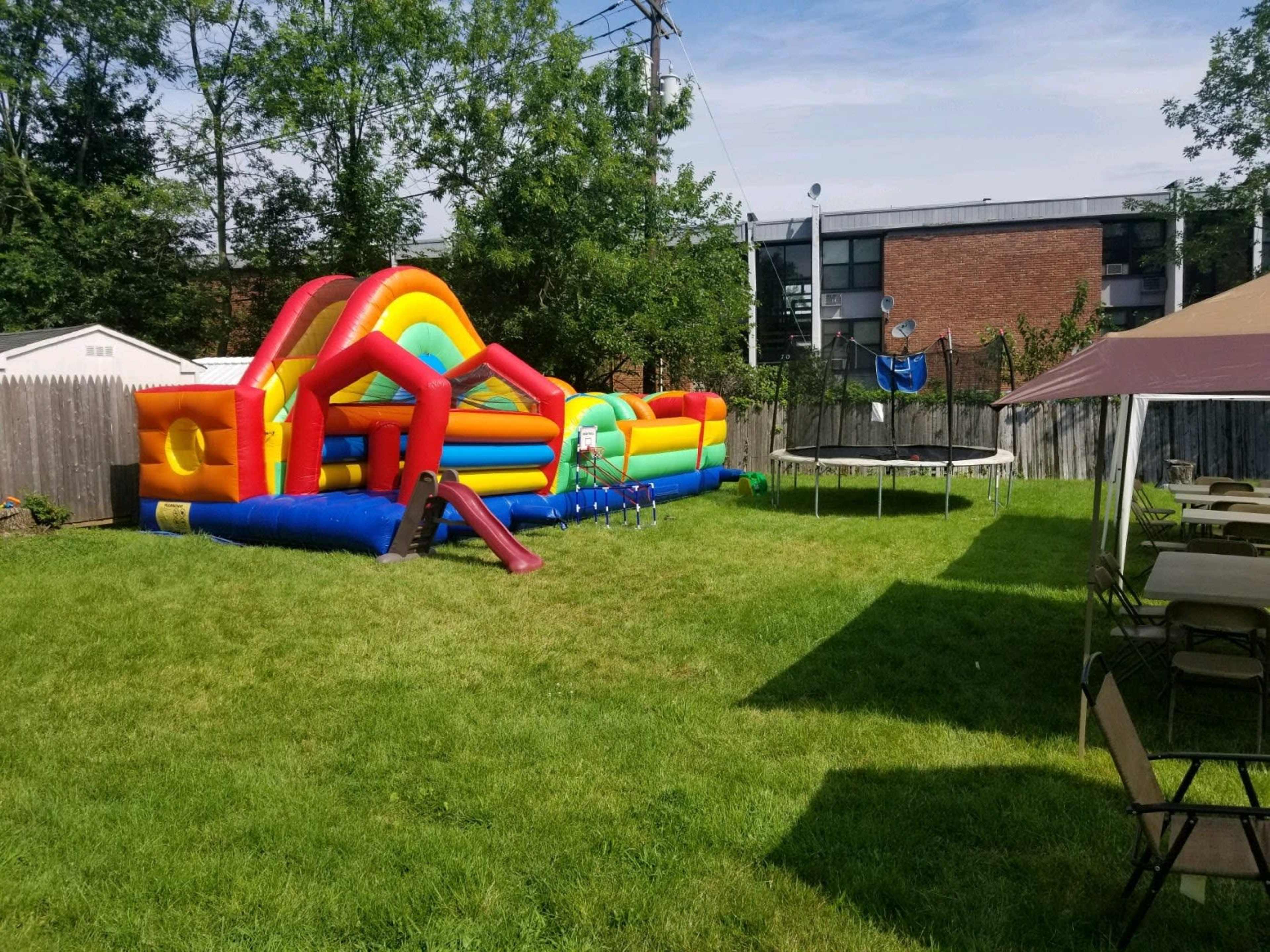 A backyard with a colorful inflatable bounce house and a trampoline near a grassy area with picnic tables set up under a canopy.