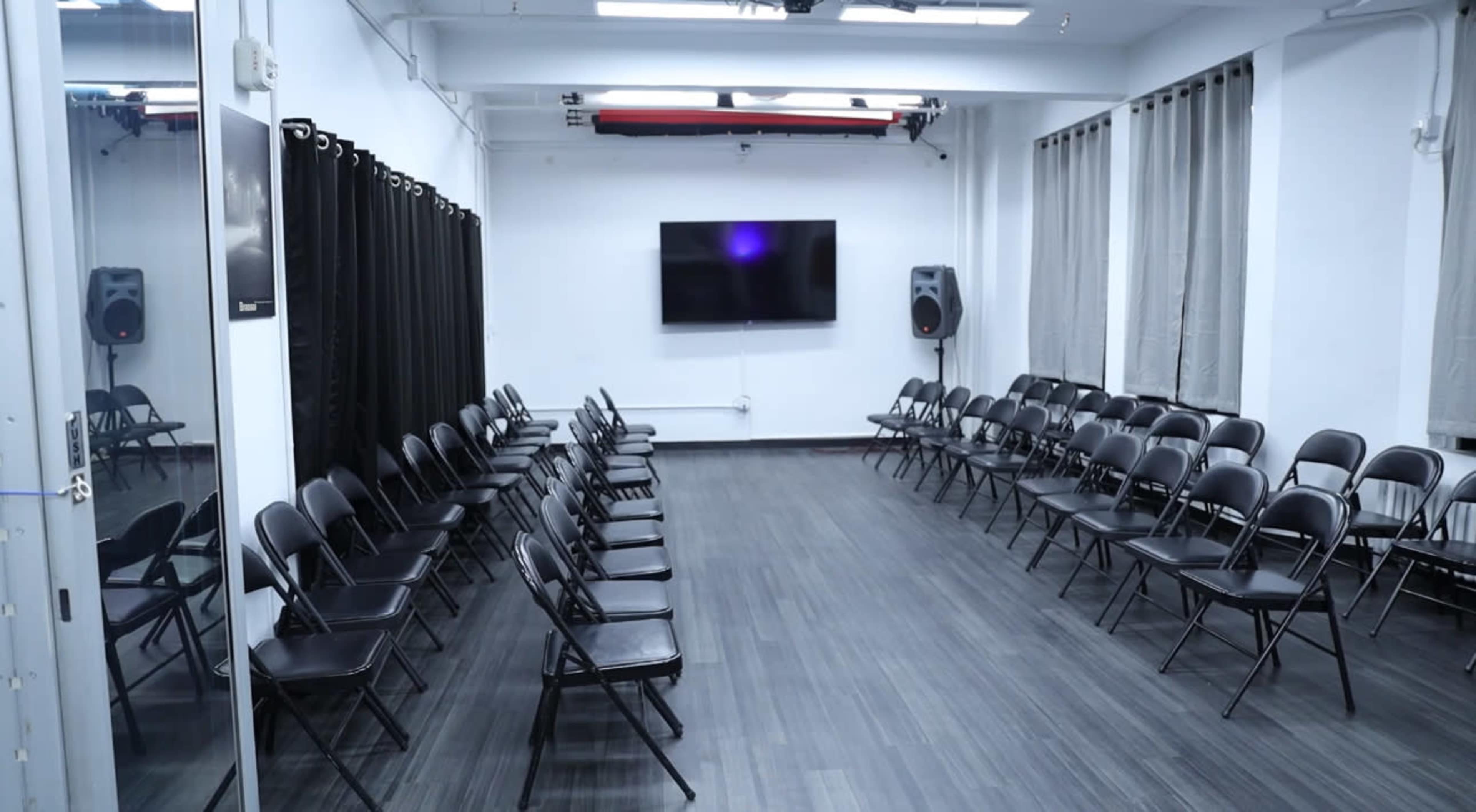 The image shows a spacious room with two rows of black folding chairs arranged facing each other, and a large screen mounted on the wall at one end.