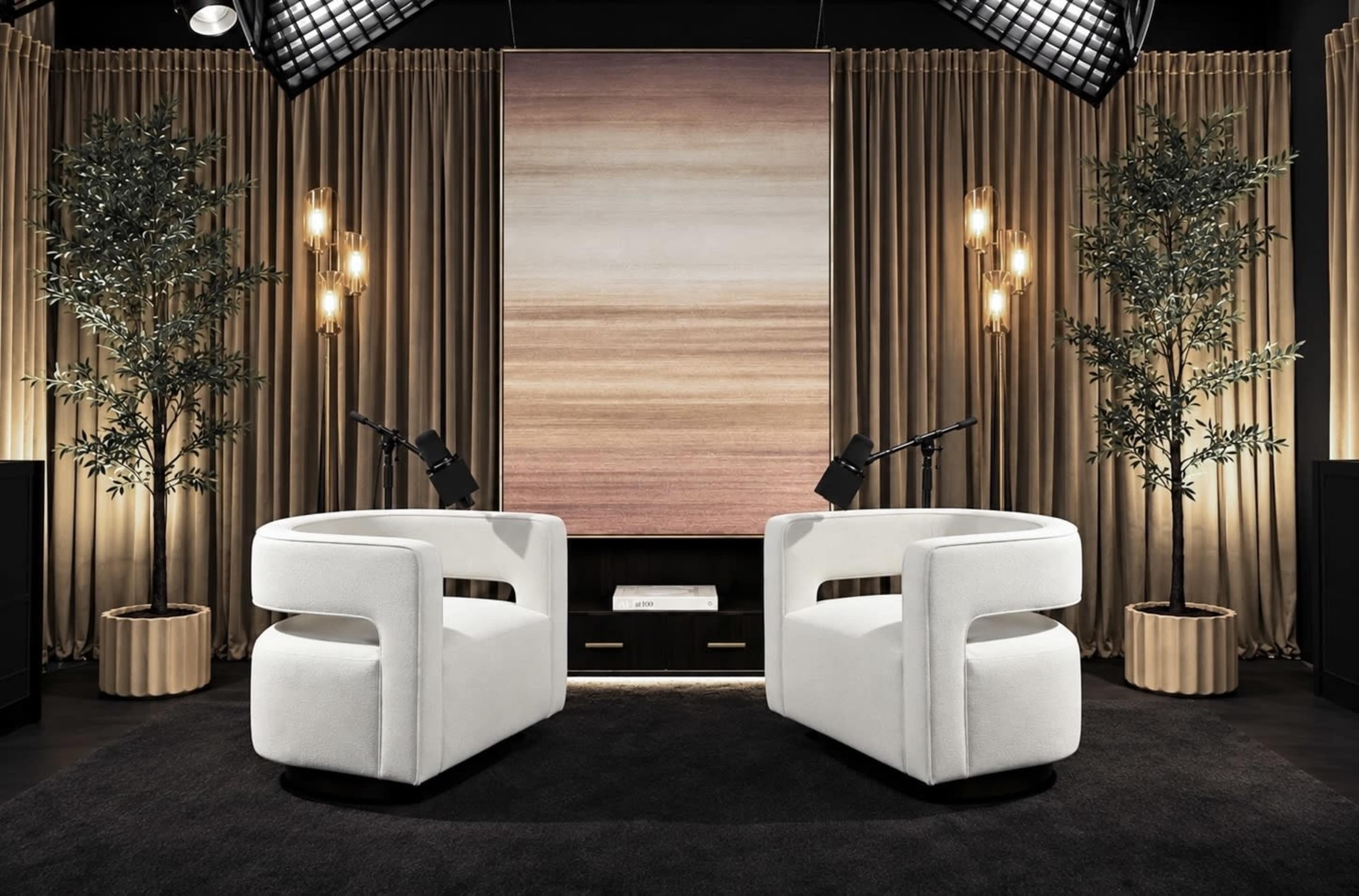 The image shows a modern studio setup with two white chairs facing each other, a large abstract painting on the wall, and decorative plants, all surrounded by warm lighting.