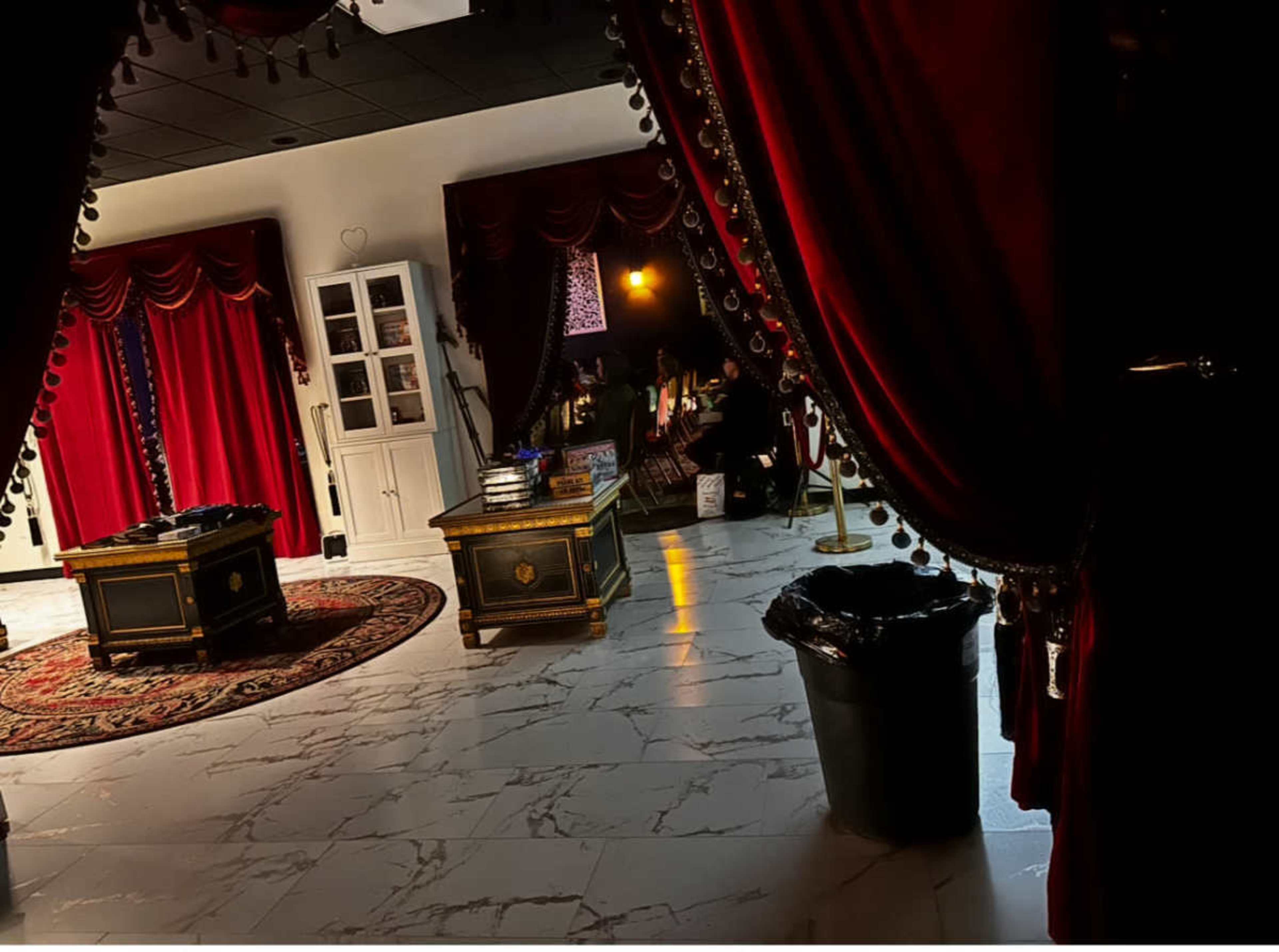 The image shows an elegantly decorated room with red drapes, marble flooring, and two ornate tables, while a trash can is visible in the foreground.